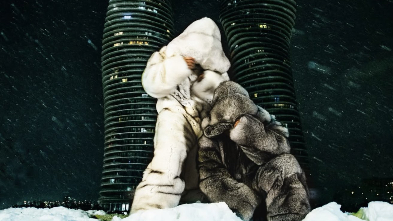 Two people in large, fluffy coats stand in front of tall, cylindrical buildings at night, with snow falling around them.