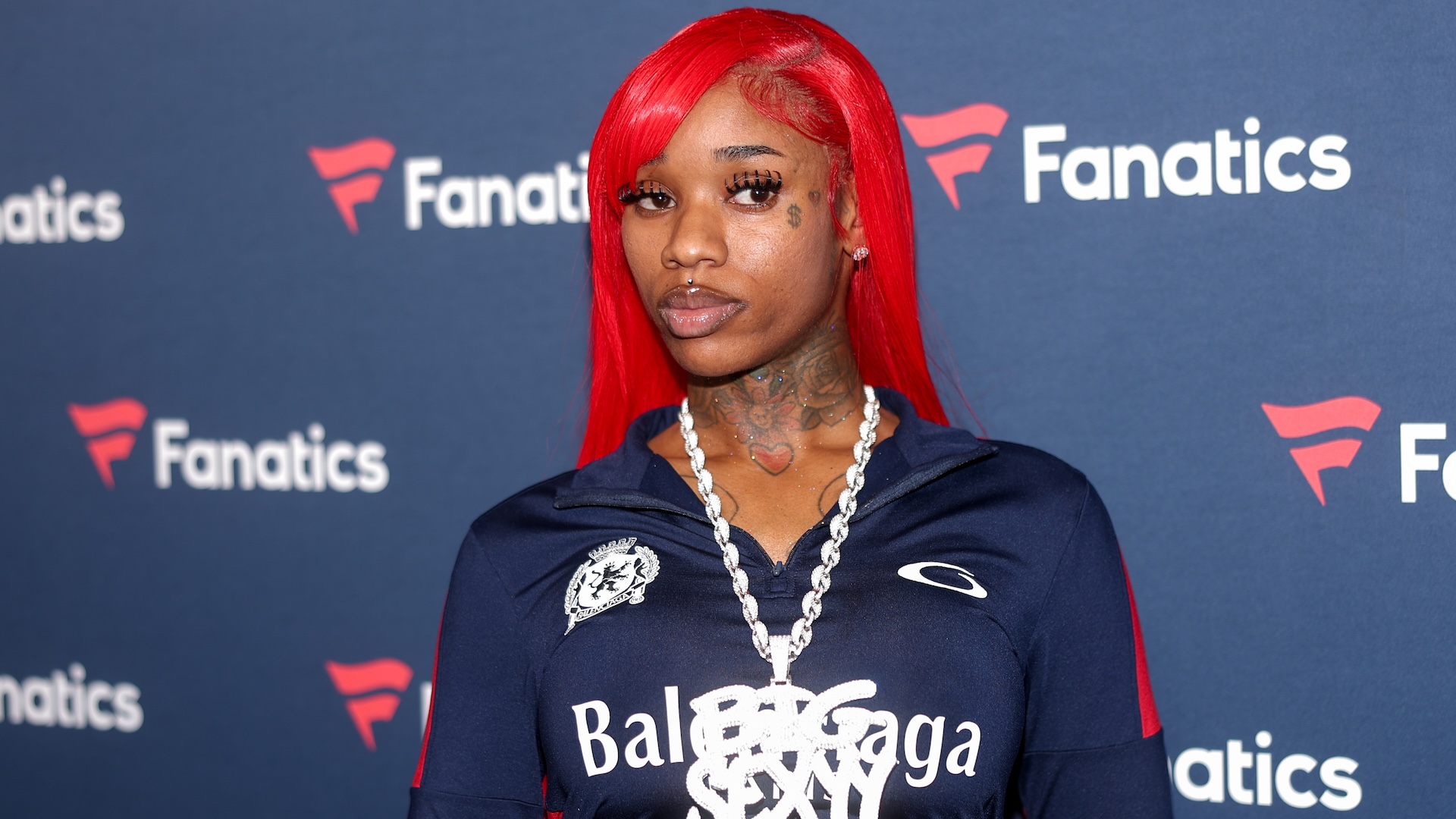 Sexyy Red with bright red hair poses in front of a Fanatics backdrop, wearing a Balenciaga top and a large chain necklace.