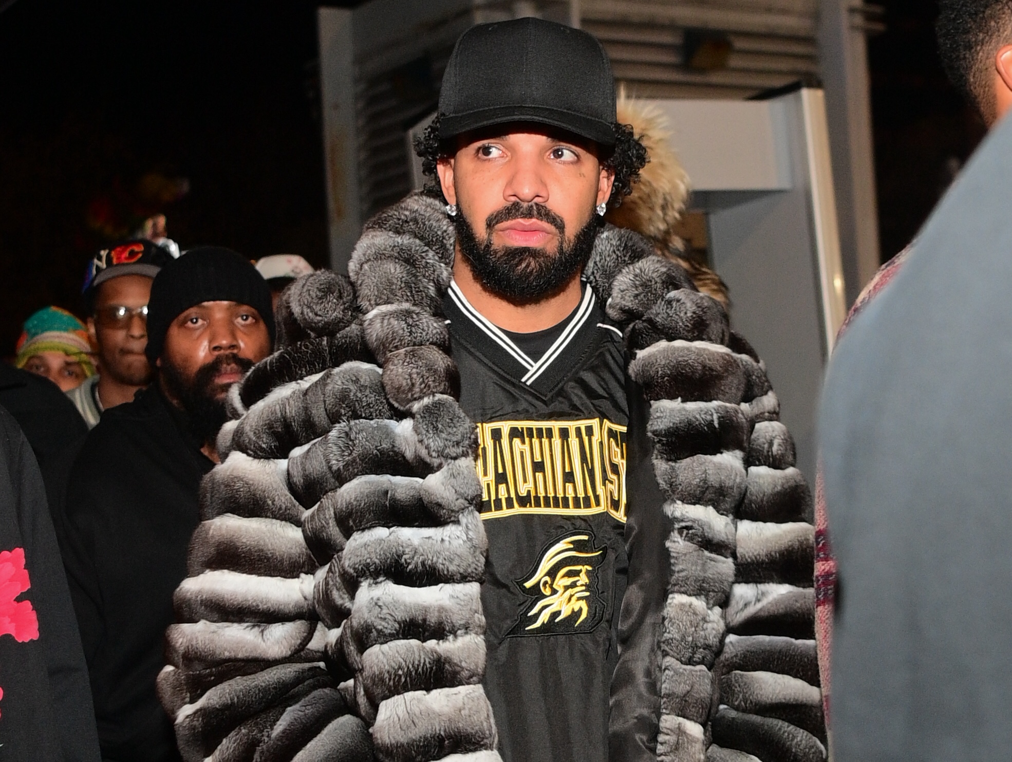 Drake wearing a fur coat and black cap, surrounded by people.