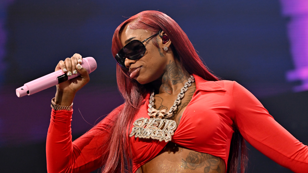 Sexyy Red with long red hair and sunglasses performs on stage, holding a pink microphone. She wears a bold necklace and a red outfit.