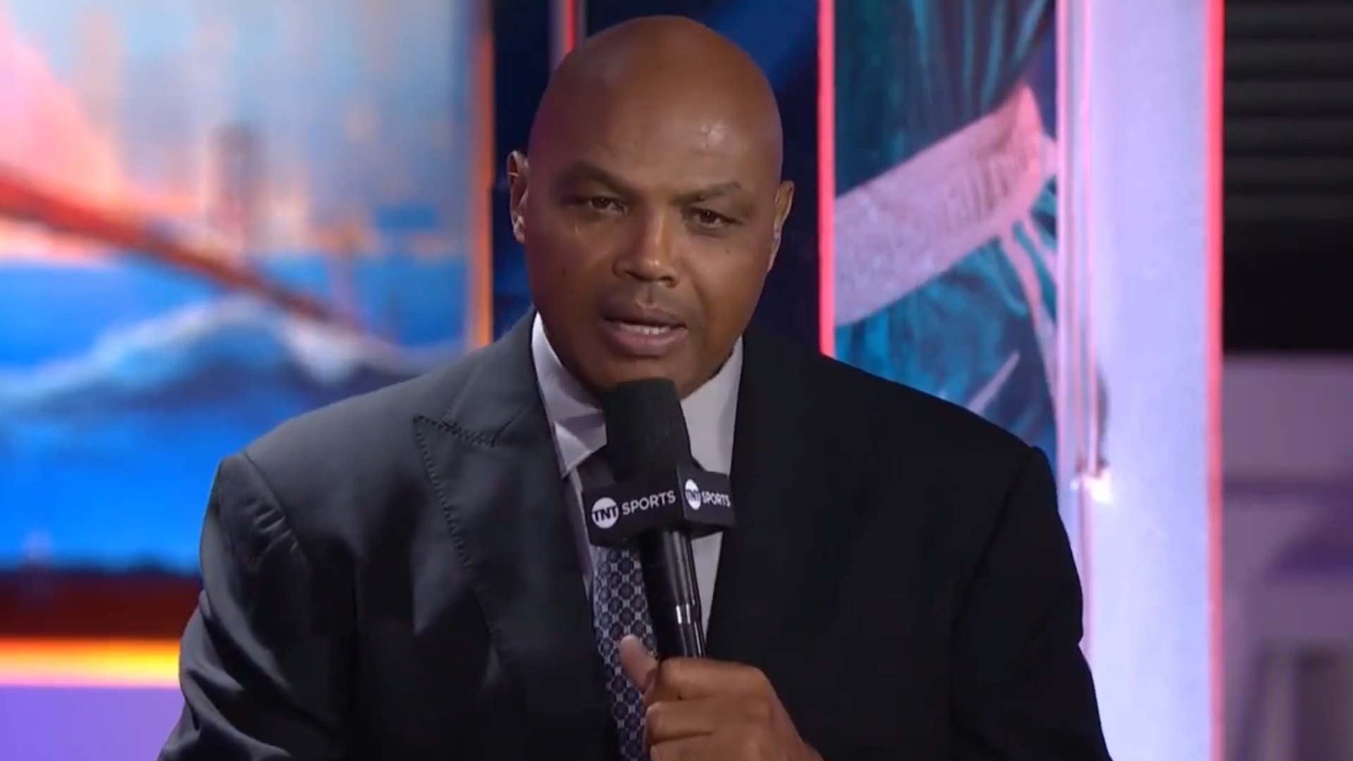 Charles Barkley speaking into a microphone on a TV set, wearing a dark suit and tie.