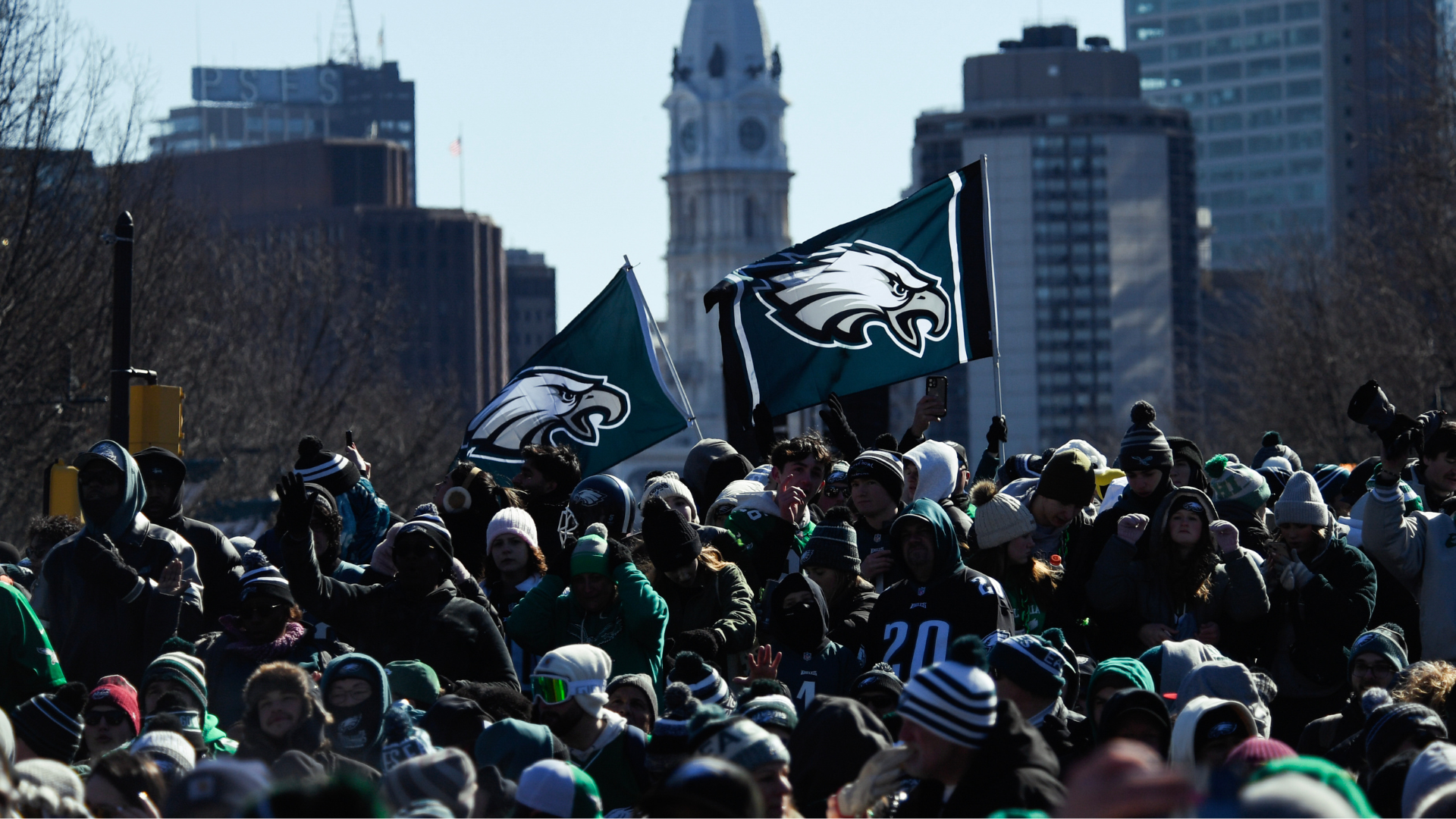 A crowd of Philadelphia Eagles fans celebrating outdoors, waving team flags. City buildings are visible in the background.