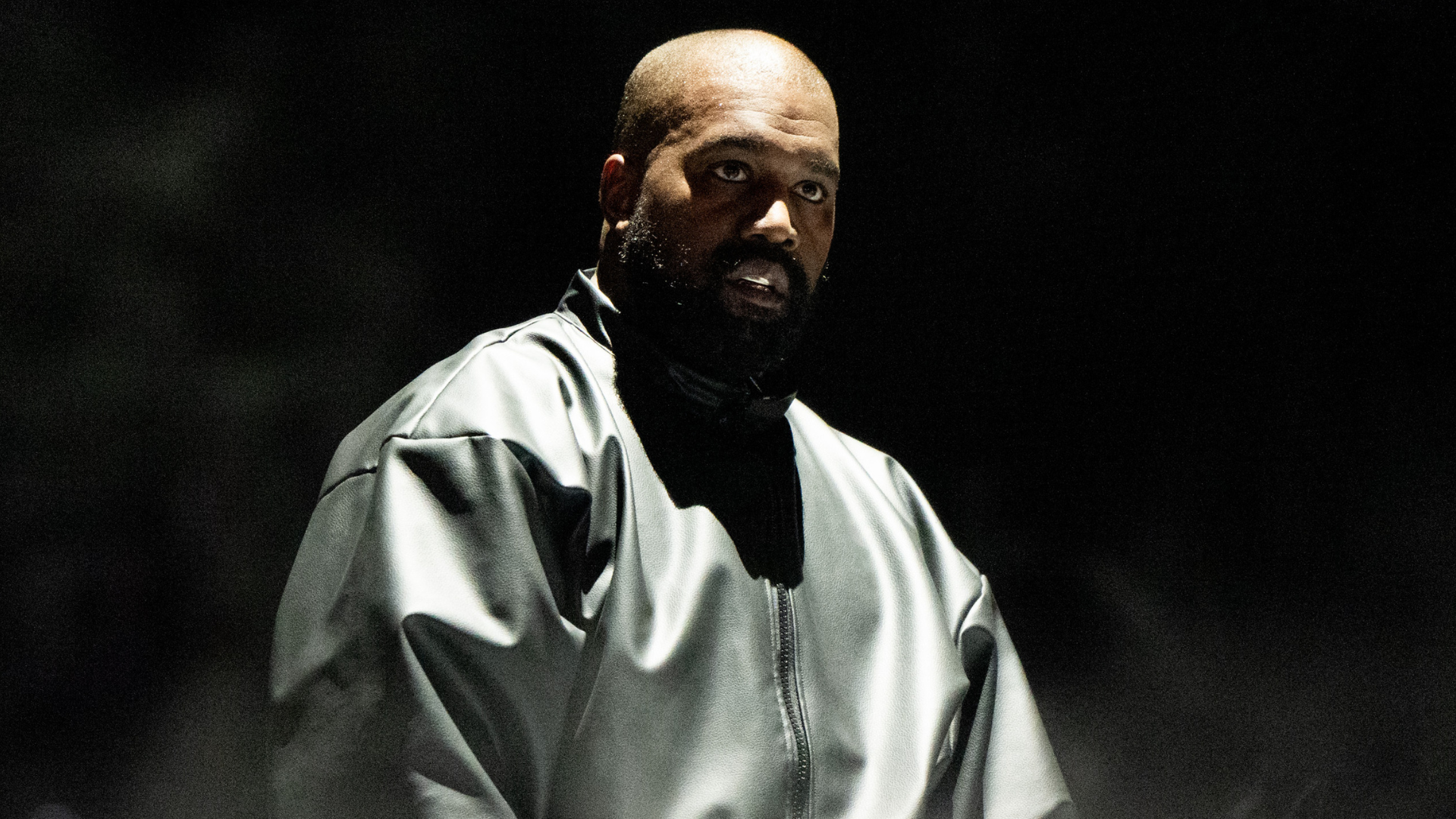 A man in a dark, oversized jacket stands against a black background, looking intently forward.