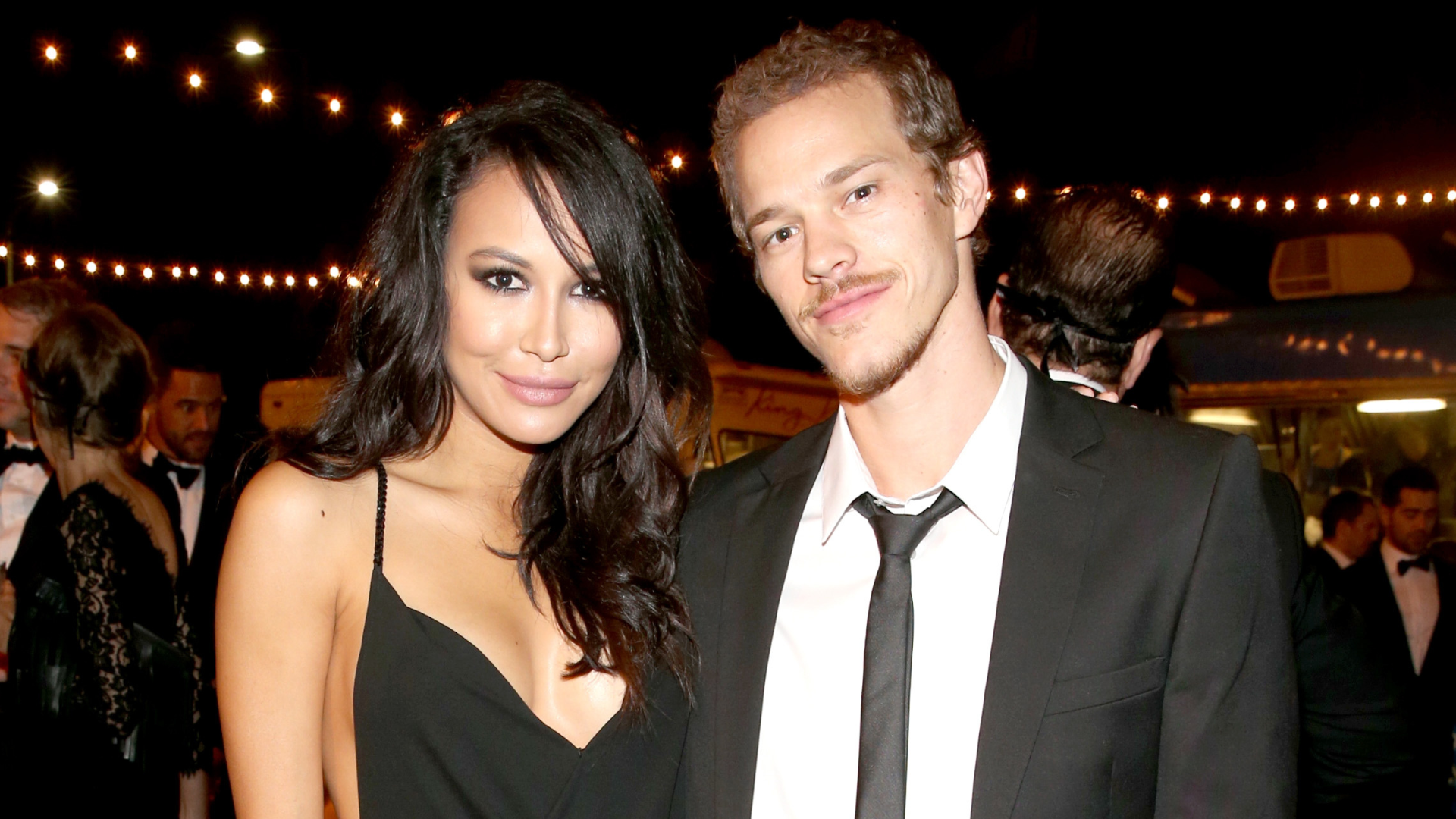 A woman in a black dress and a man in a suit pose together at an event with string lights in the background.