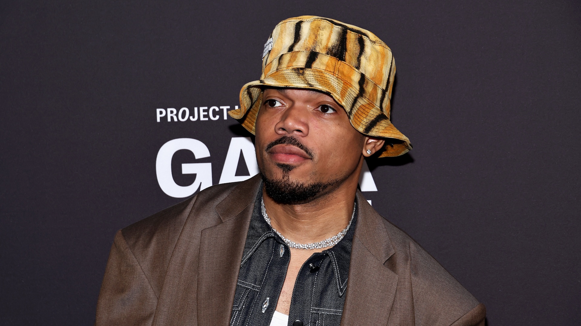Chance the Rapper wearing a tiger-striped bucket hat and brown jacket at an event, standing against a dark background.