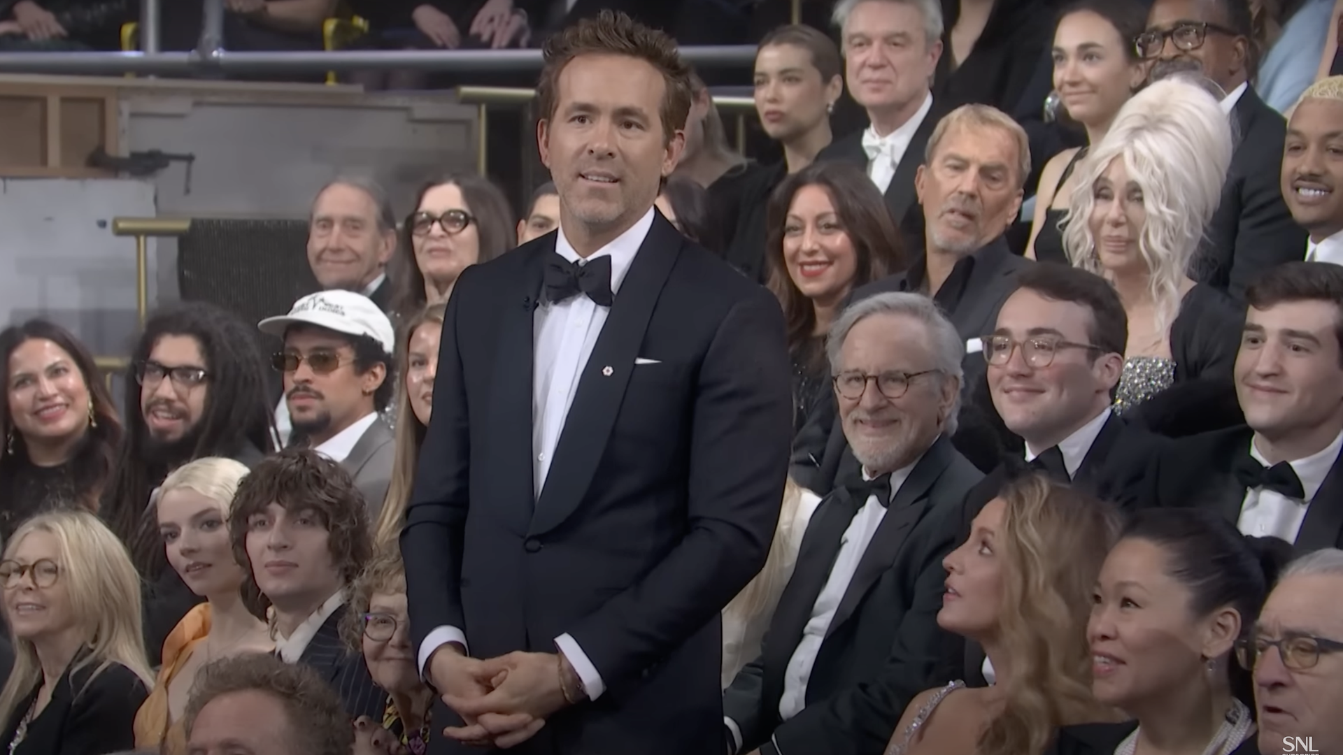 Ryan Reynolds stands among a crowd in formal attire. Steven Spielberg and other attendees are seated around him, all smiling.