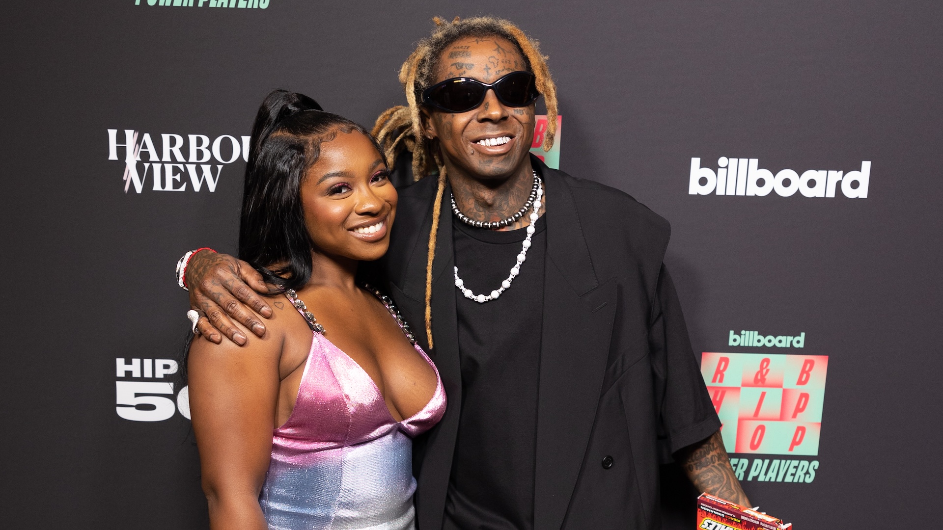 Lil Wayne and Reginae Carter pose together at an event, with Lil Wayne wearing sunglasses and a black outfit, and Reginae in a colorful dress.