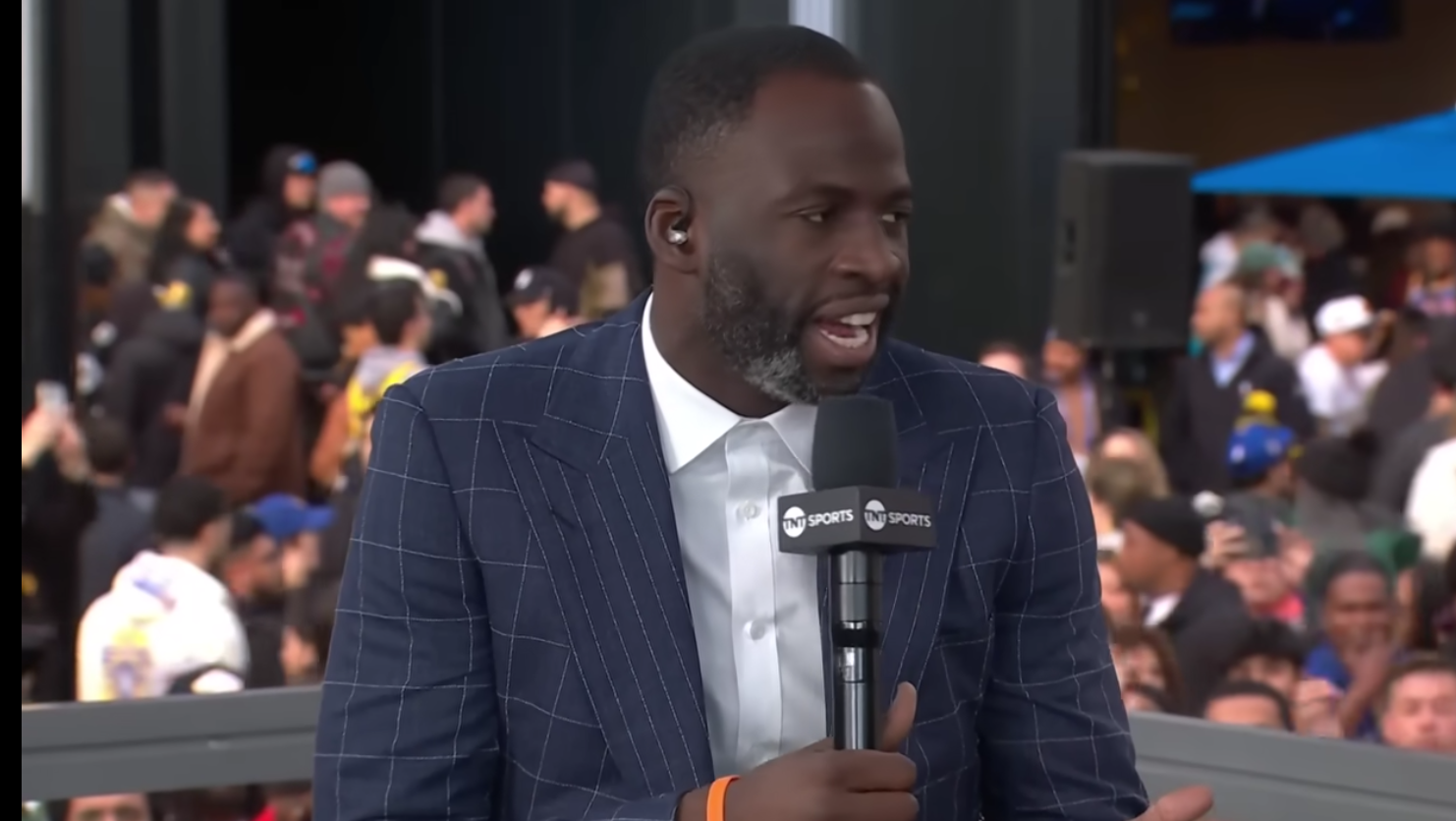 A man in a pinstripe suit speaks into a microphone at an event, with a crowd of people in the background.