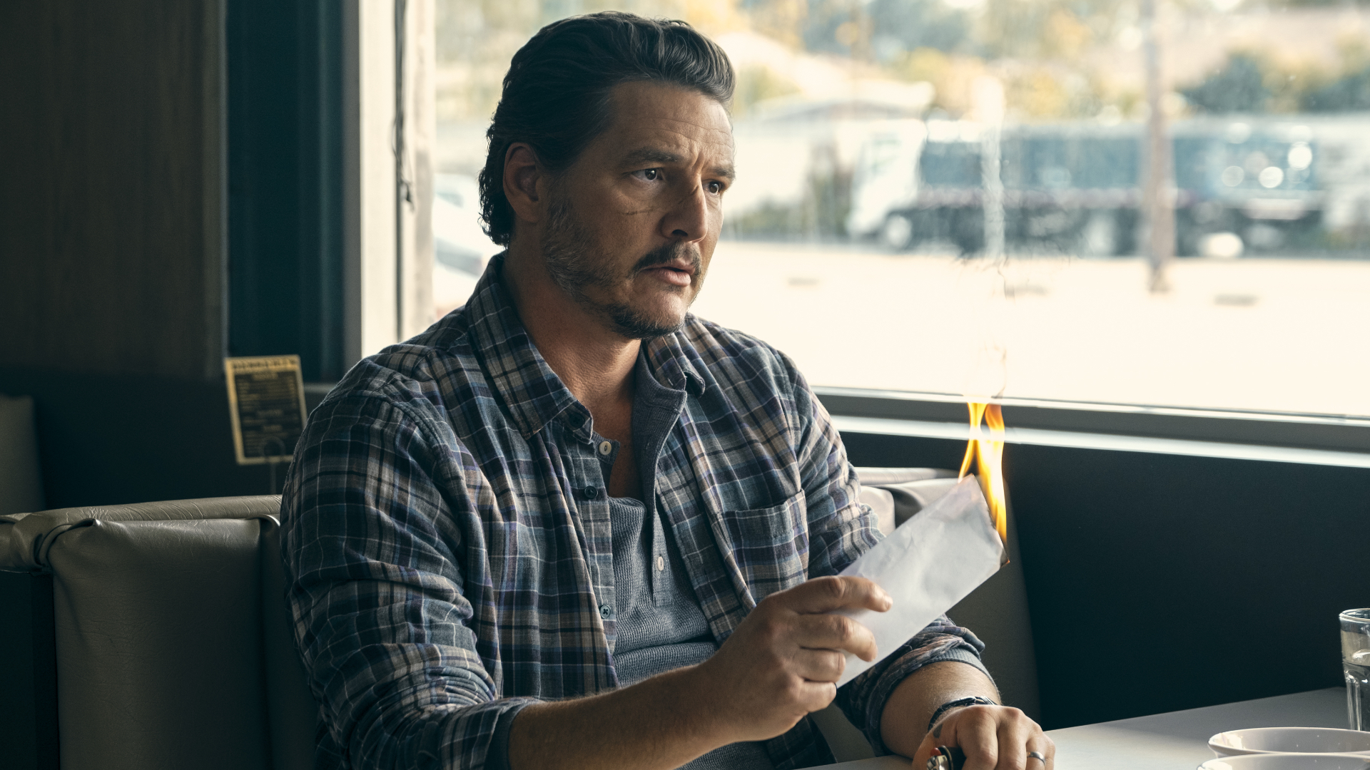 Pedro Pascal in a plaid shirt sits at a diner table, holding a burning paper. Sunlight streams through the window behind him.