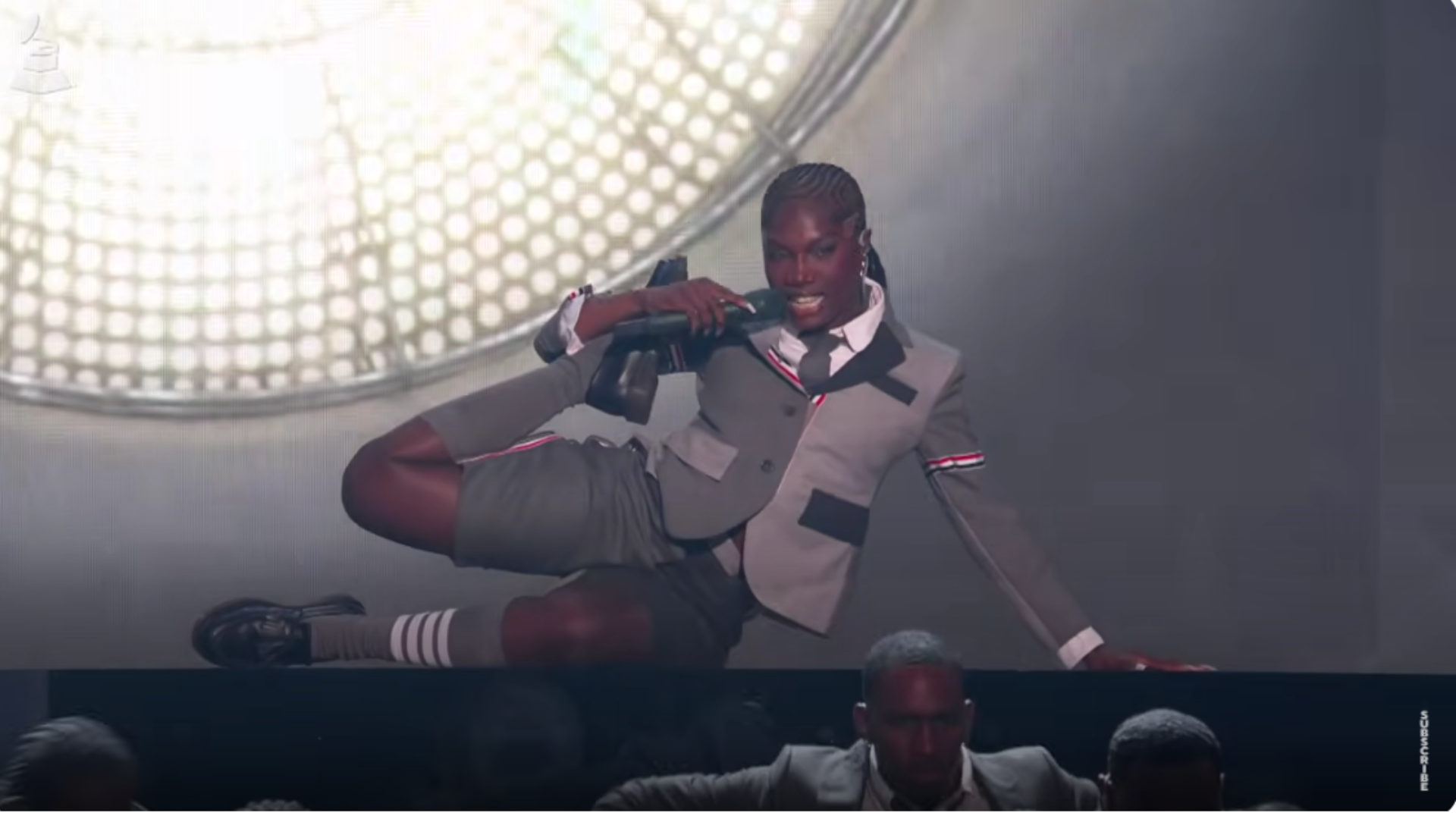Performer in a gray suit and shorts, lying on a stage with a microphone, in front of a circular light pattern.