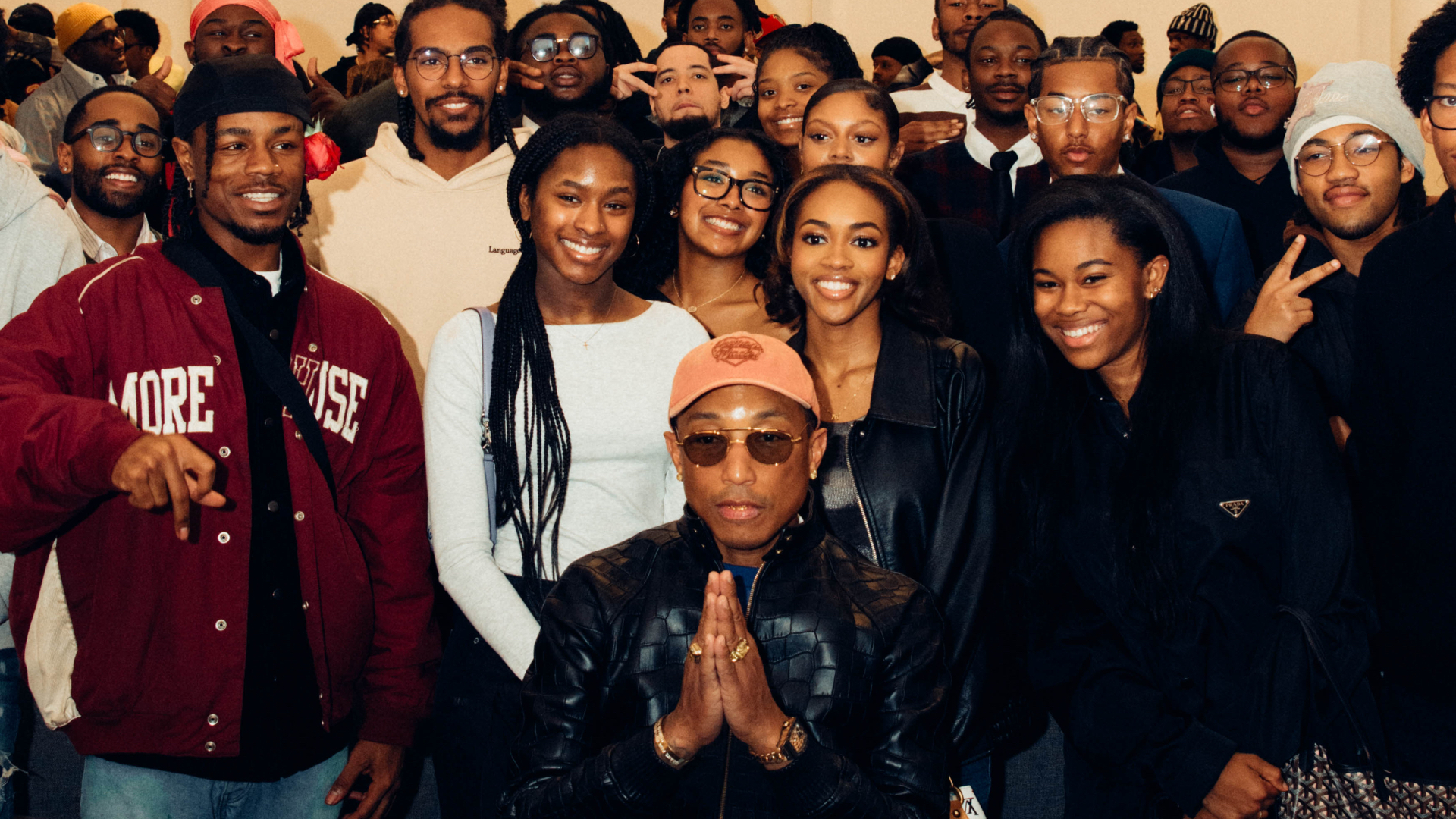 Pharrell Williams in sunglasses and a cap, posing with a group of smiling people.