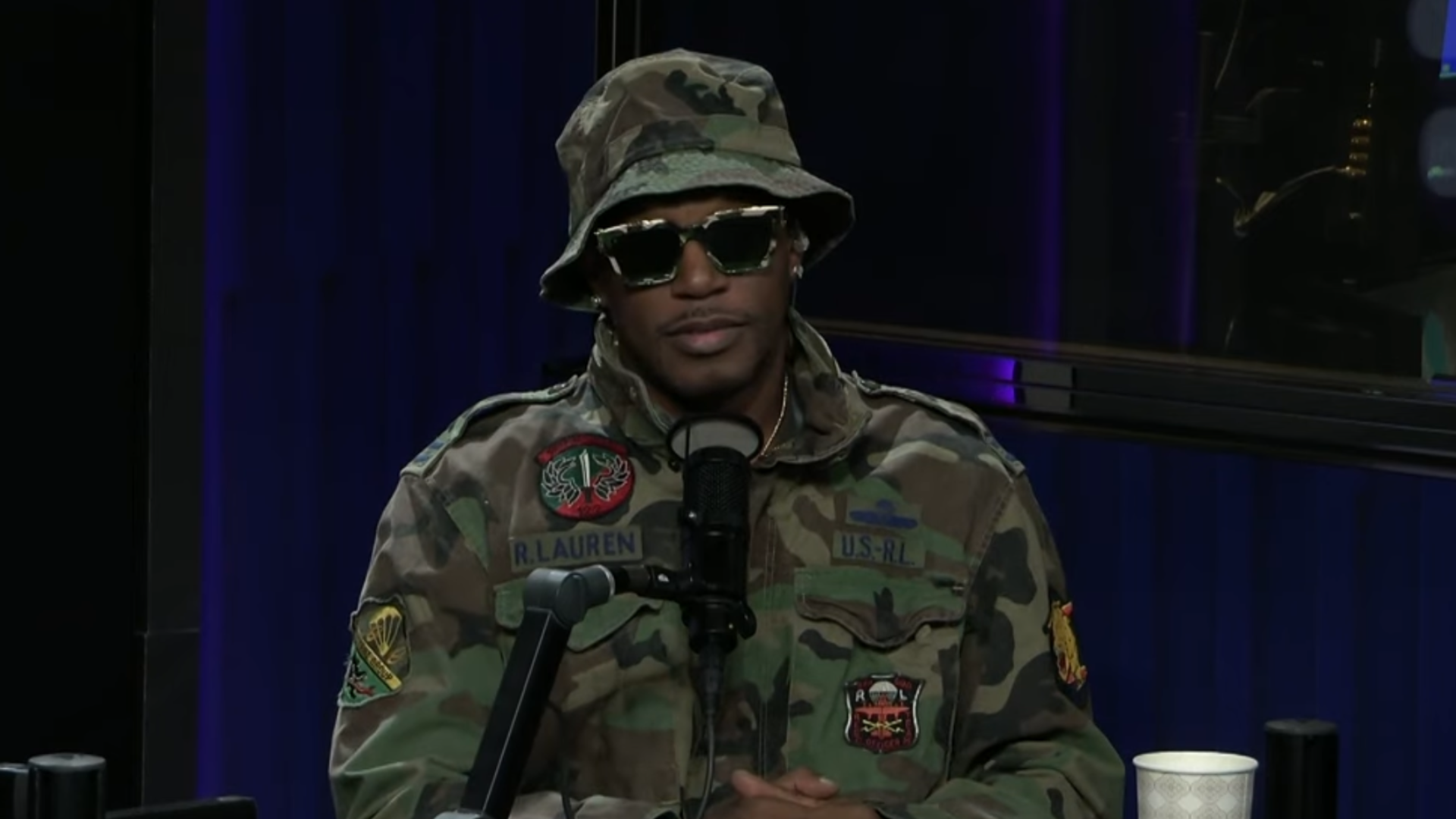 A person in a camo jacket and hat, wearing sunglasses, sits at a microphone in a studio setting.