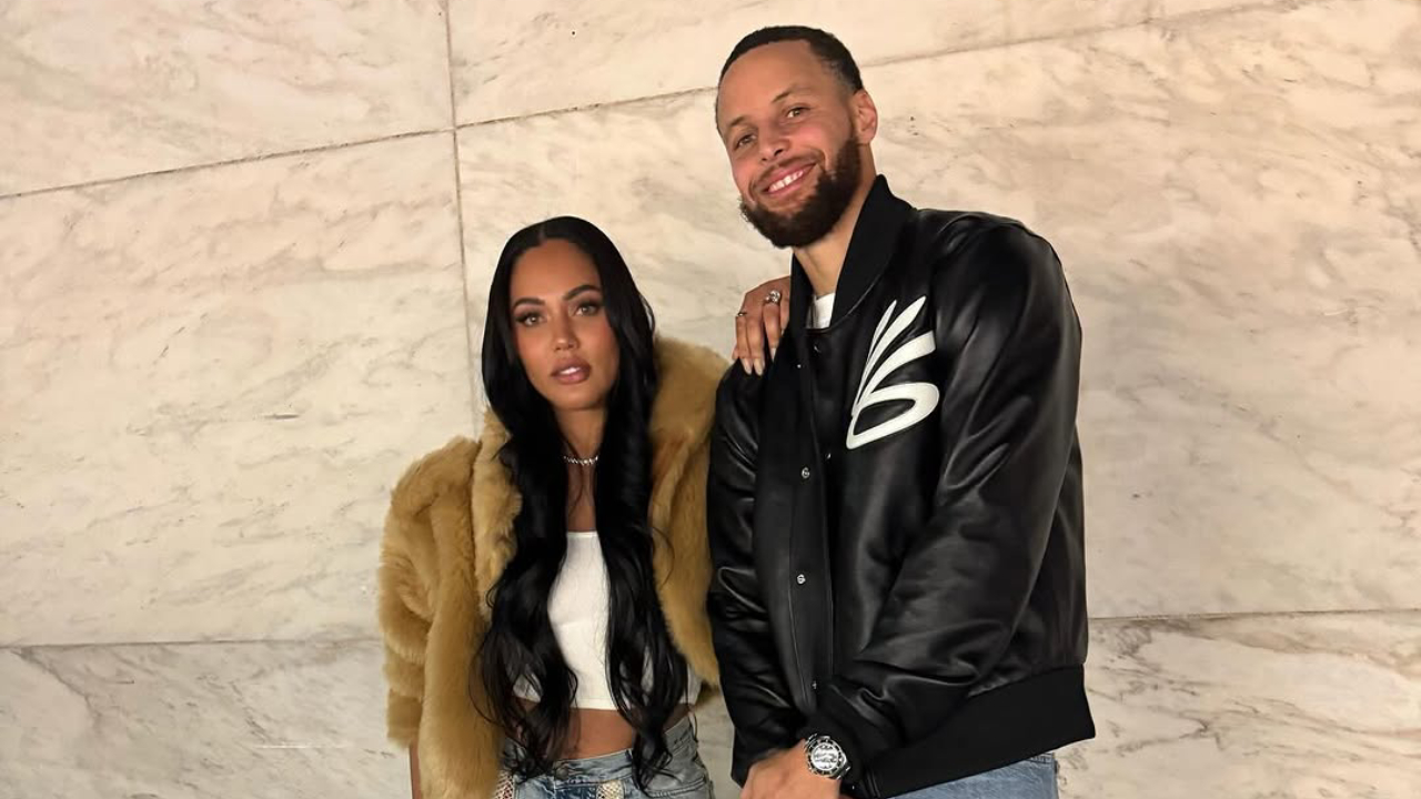 Ayesha and Stephen Curry posing together. Stephen is wearing a black jacket while Ayesha dons a fur coat. They stand against a marble wall.