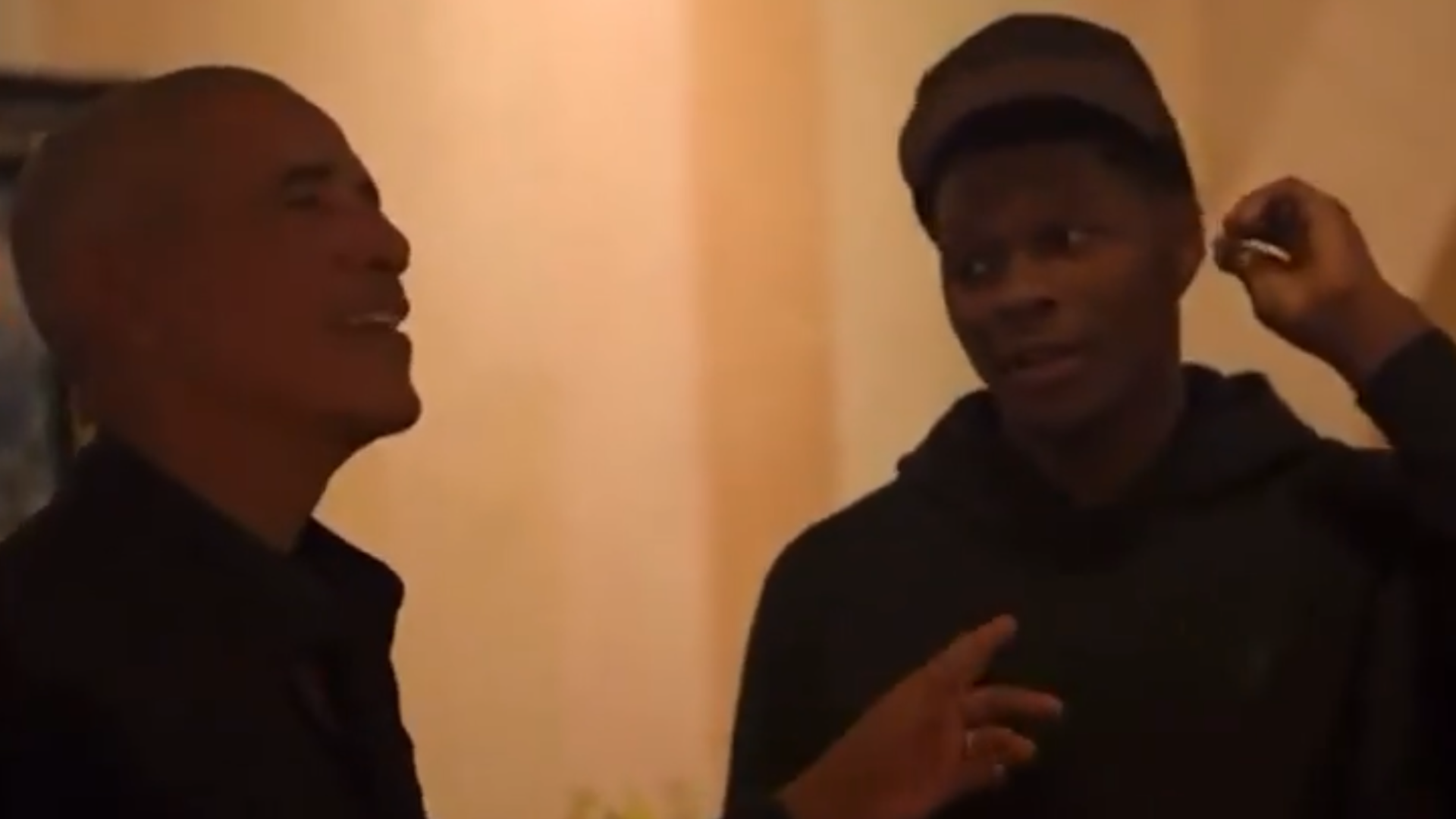 Two people are engaged in conversation indoors, one wearing a dark jacket and the other a hoodie and cap.