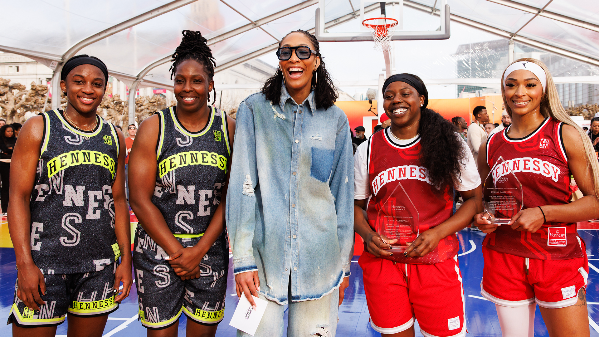 A group of women, including a tall woman in sunglasses and denim, stand on a basketball court. Some wear "Hennessy" jerseys.