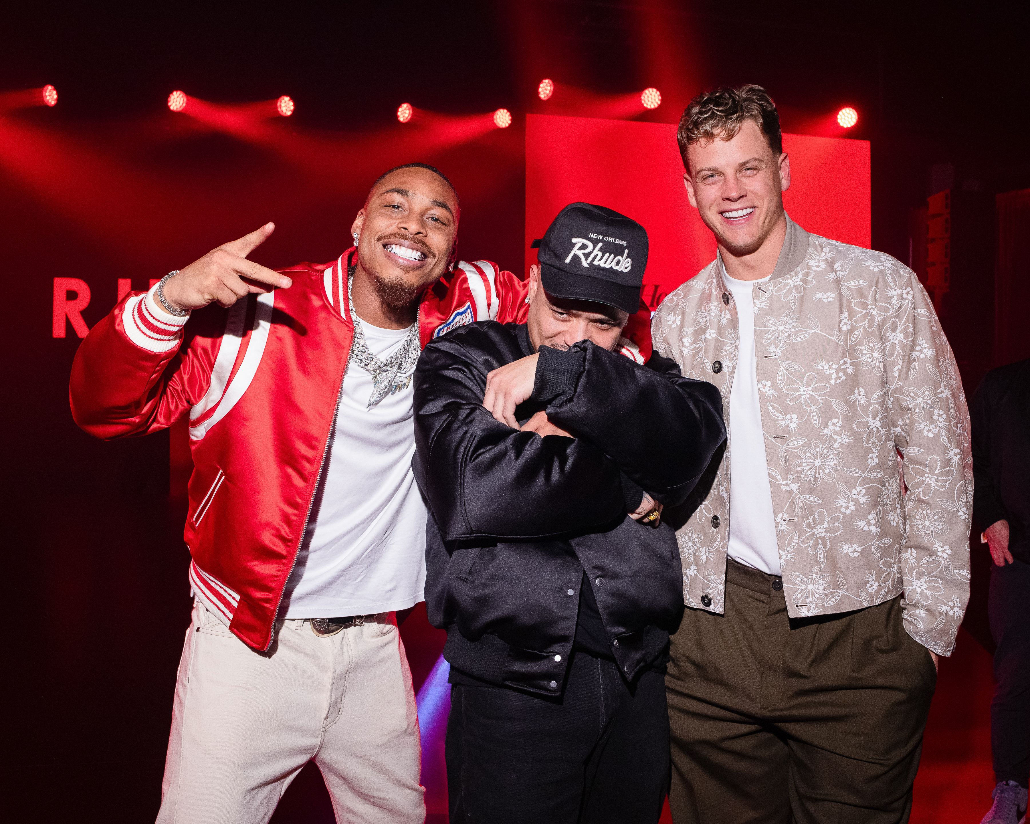 Three men posing together at an event, one wearing a red jacket, another in a black Rhude cap, and the third in a floral jacket.
