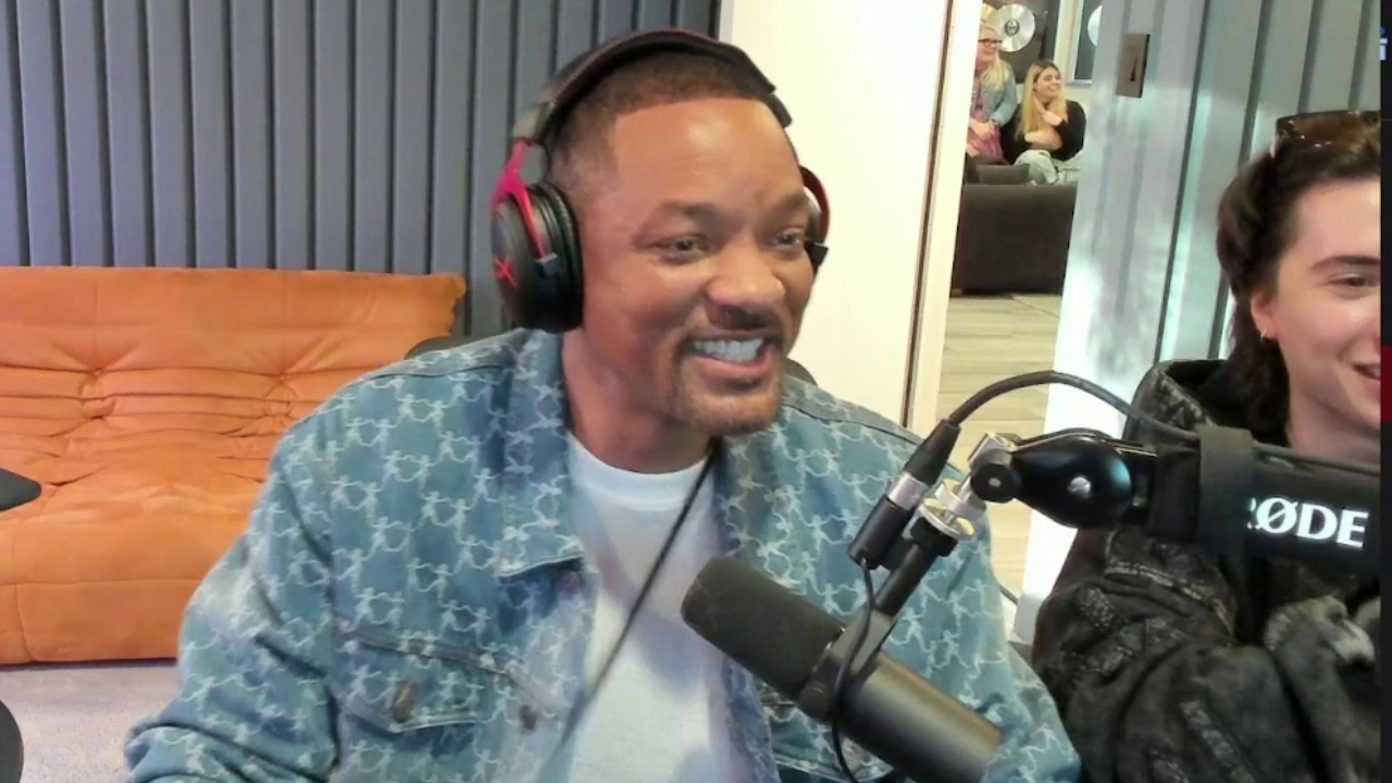 Will Smith, wearing headphones and a blue jacket, smiles while speaking into a microphone.