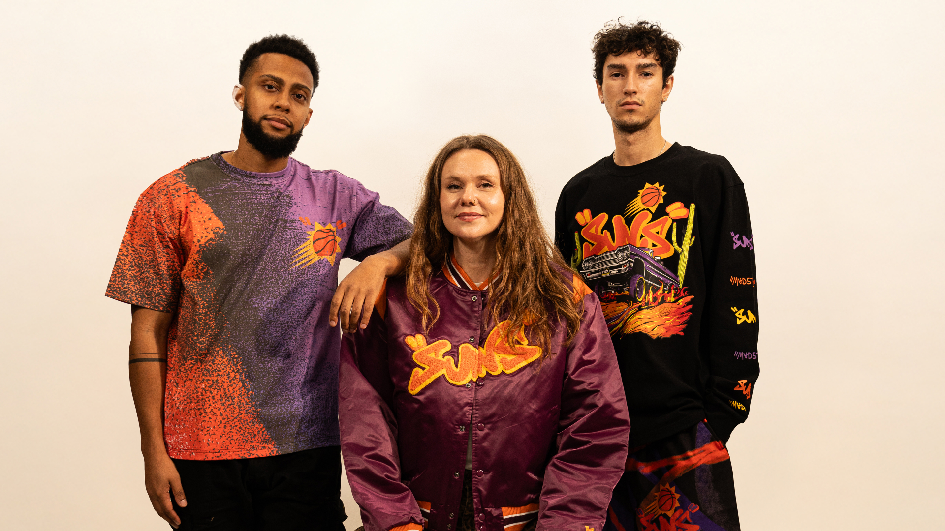 Three people wearing Phoenix Suns-themed clothing, including a purple jacket and colorful shirts, standing against a plain background.