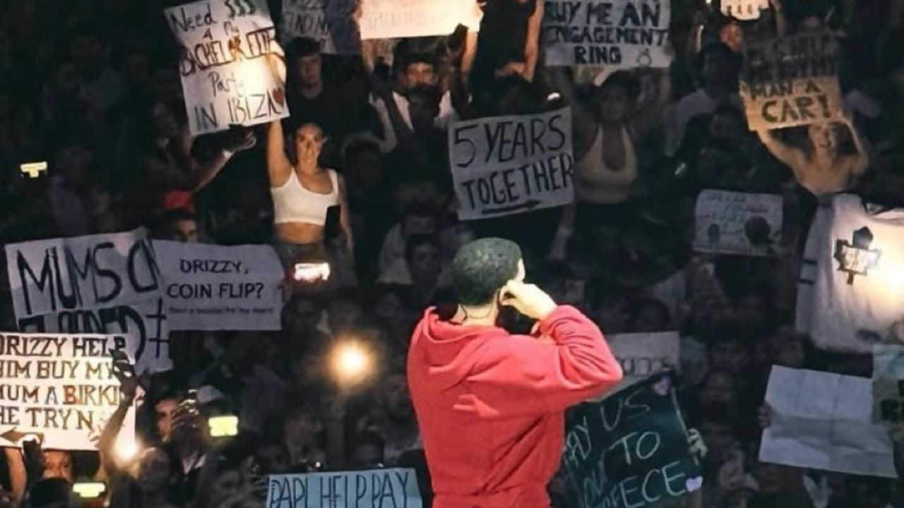 Drake on stage in a red outfit, facing a crowd holding various signs with messages like "5 years together" and "Drizzy help."