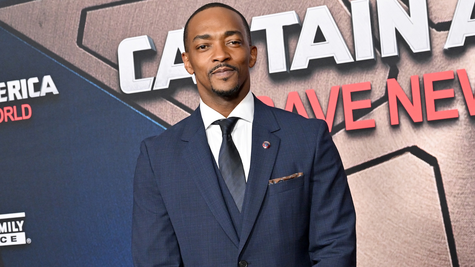 Anthony Mackie in a suit at a "Captain America" event, standing in front of a promotional backdrop.