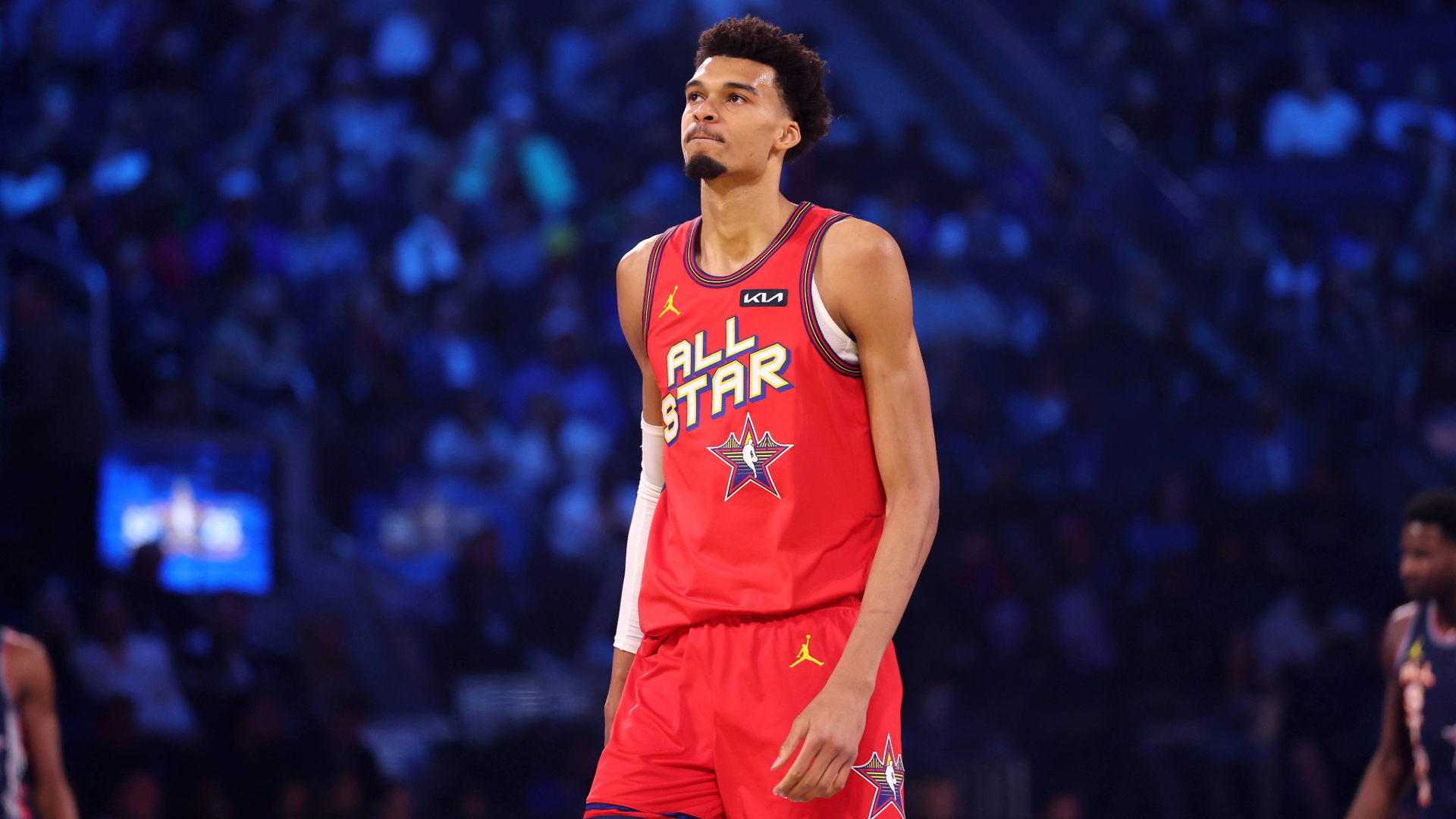 A basketball player in a red "All Star" jersey stands on the court, with a blurred audience in the background.