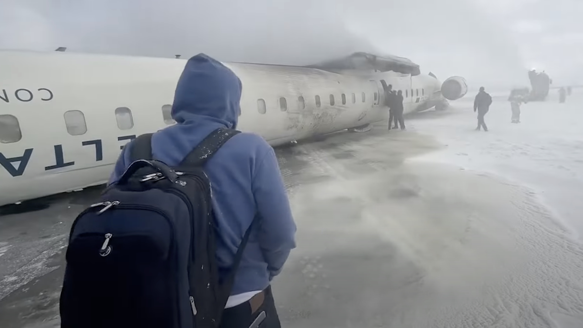 A person in a blue hoodie and backpack walks toward a crashed airplane on a snowy runway, with smoke and people nearby.