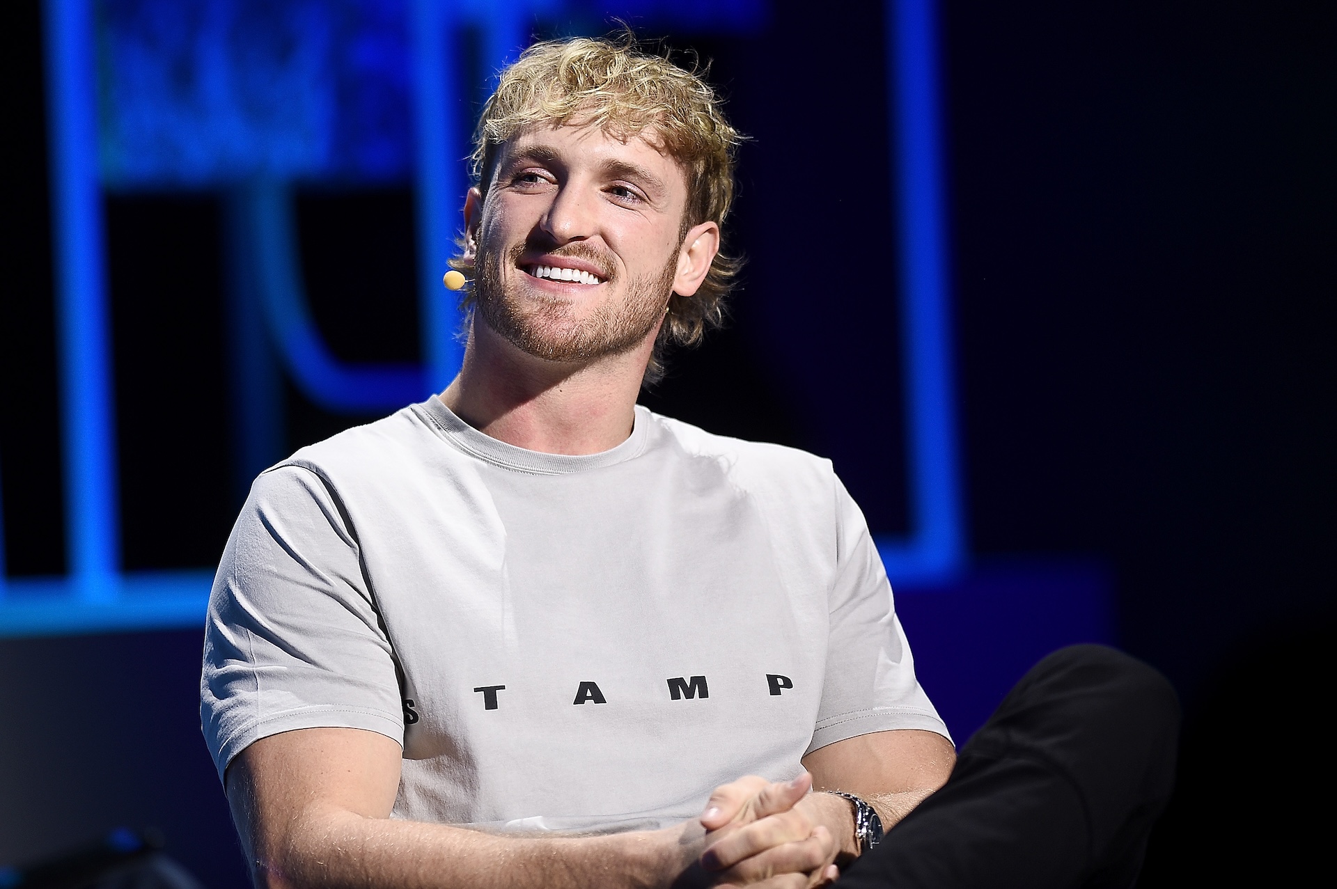 Logan Paul, wearing a light gray T-shirt, smiles while seated on stage with a microphone headset.