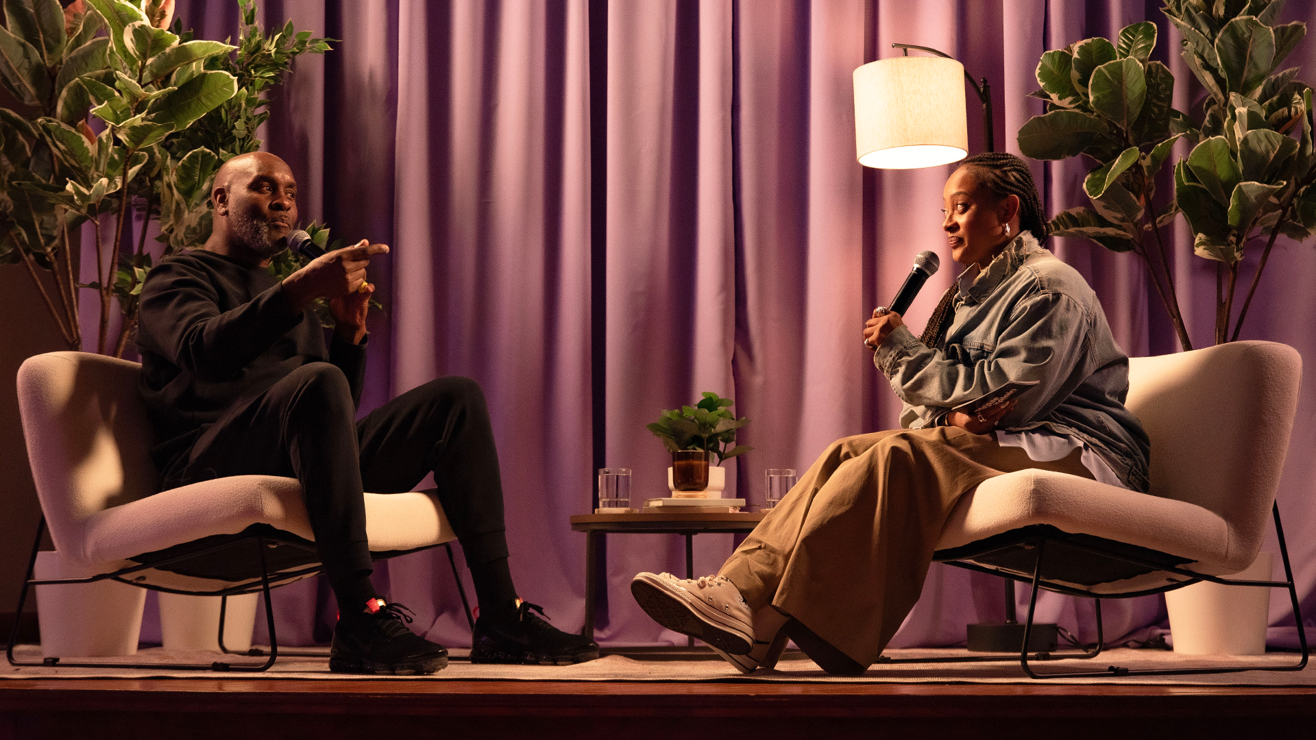 Two people sitting on stage in conversation, one holding a microphone. The setting has a purple curtain, plants, and soft lighting.