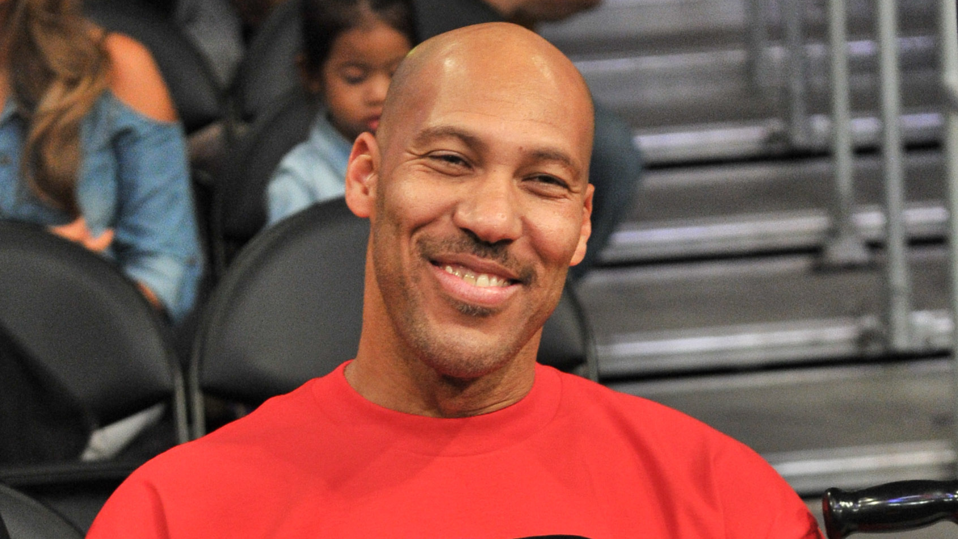 A man in a red shirt smiling while seated in an arena, with people in the background.