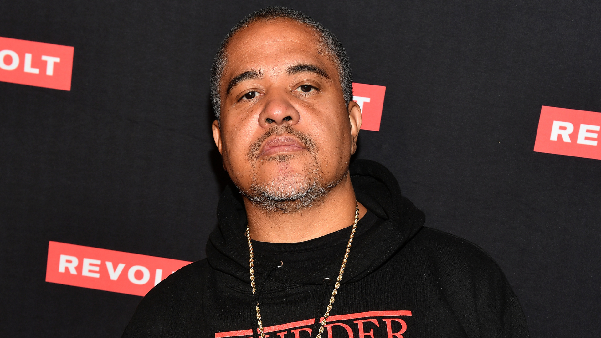 Man in a black hoodie with a chain necklace, standing against a backdrop with "REVOLT" logos.