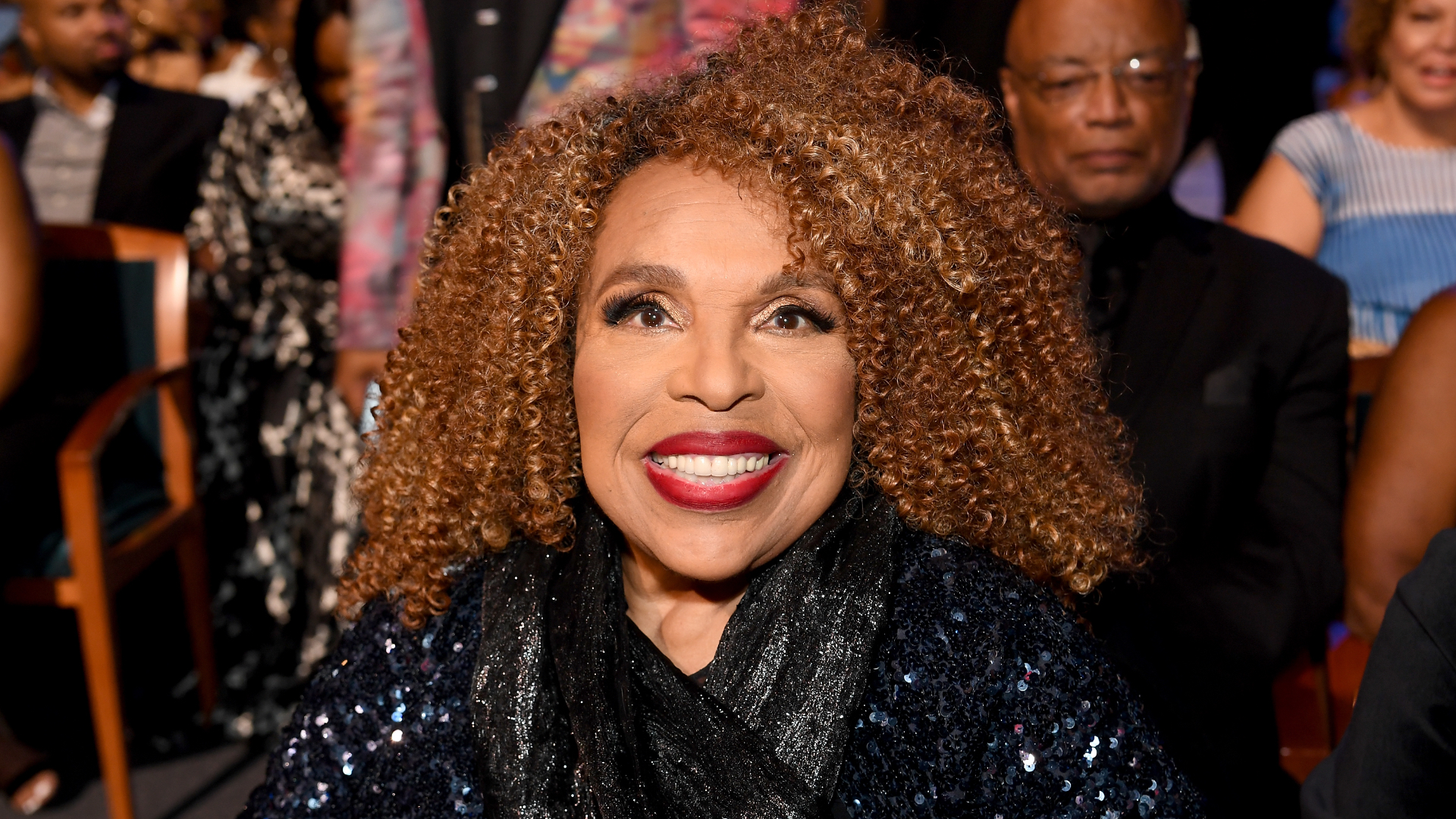 A woman with curly hair and a sparkly outfit smiles at an event, surrounded by other attendees.