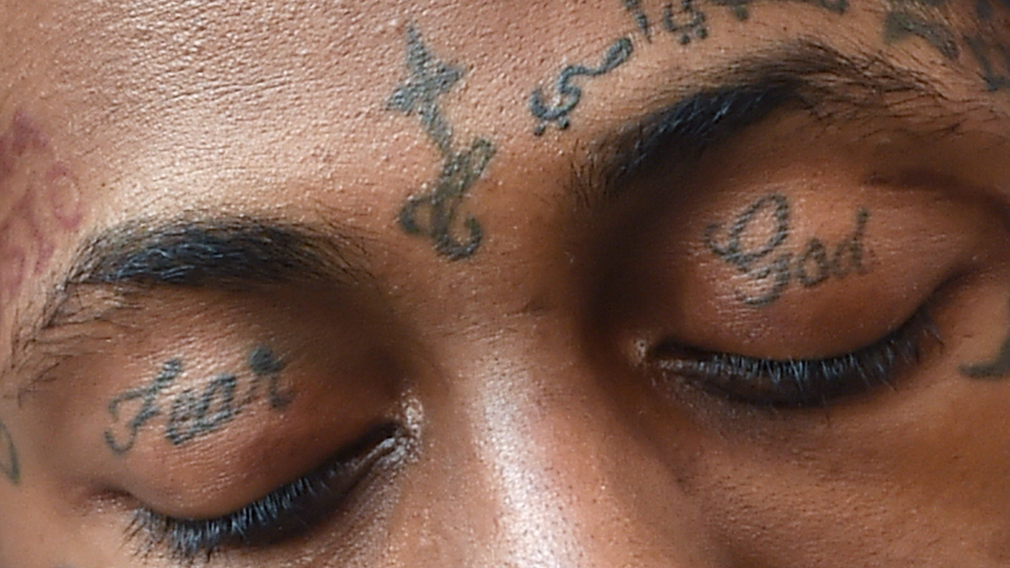 Close-up of a person's closed eyes with tattoos above the eyebrows. The tattoos read "Fear" and "God" on the eyelids.