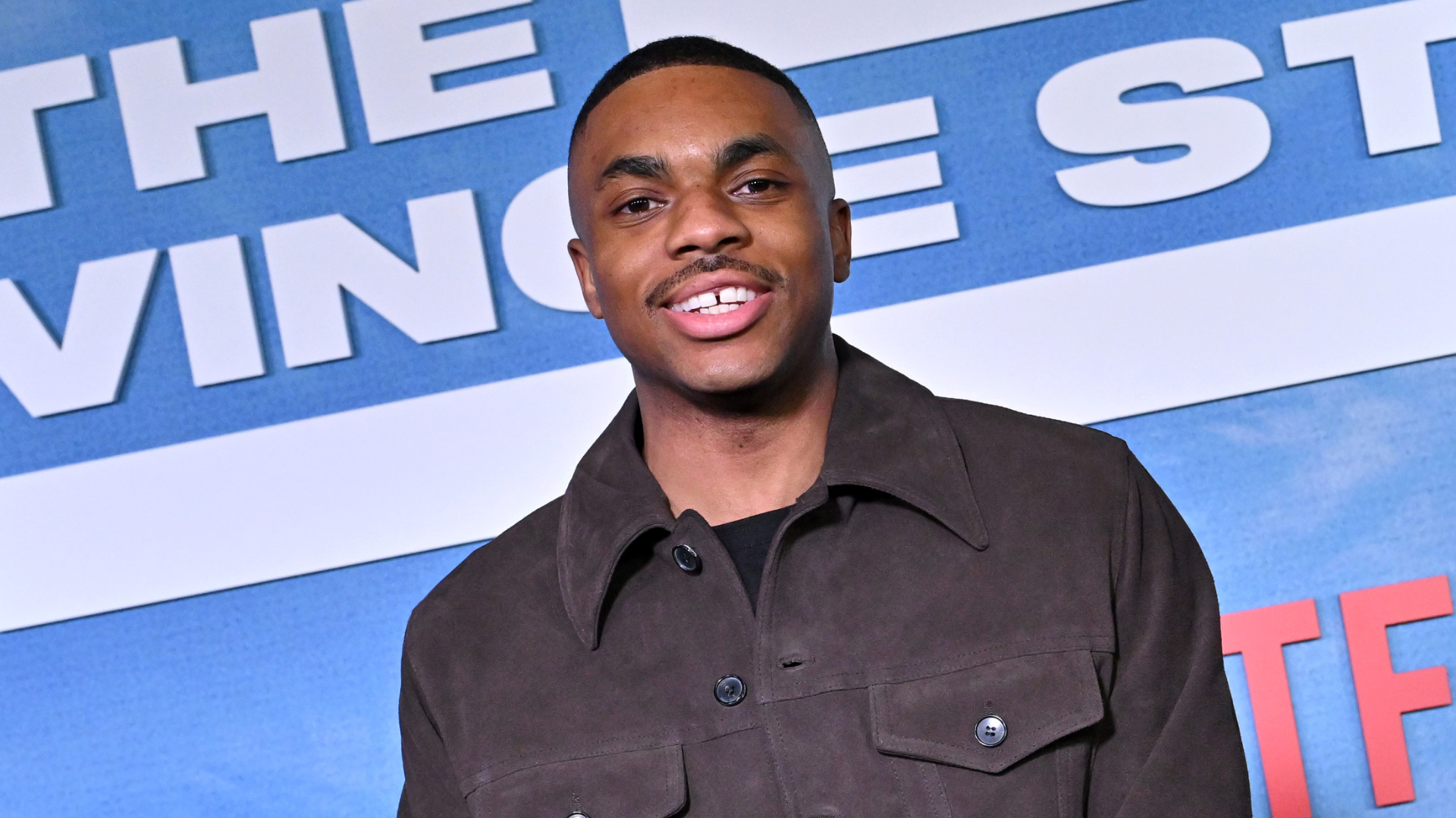 Vince Staples at an event, wearing a brown jacket, smiling in front of a backdrop with text.