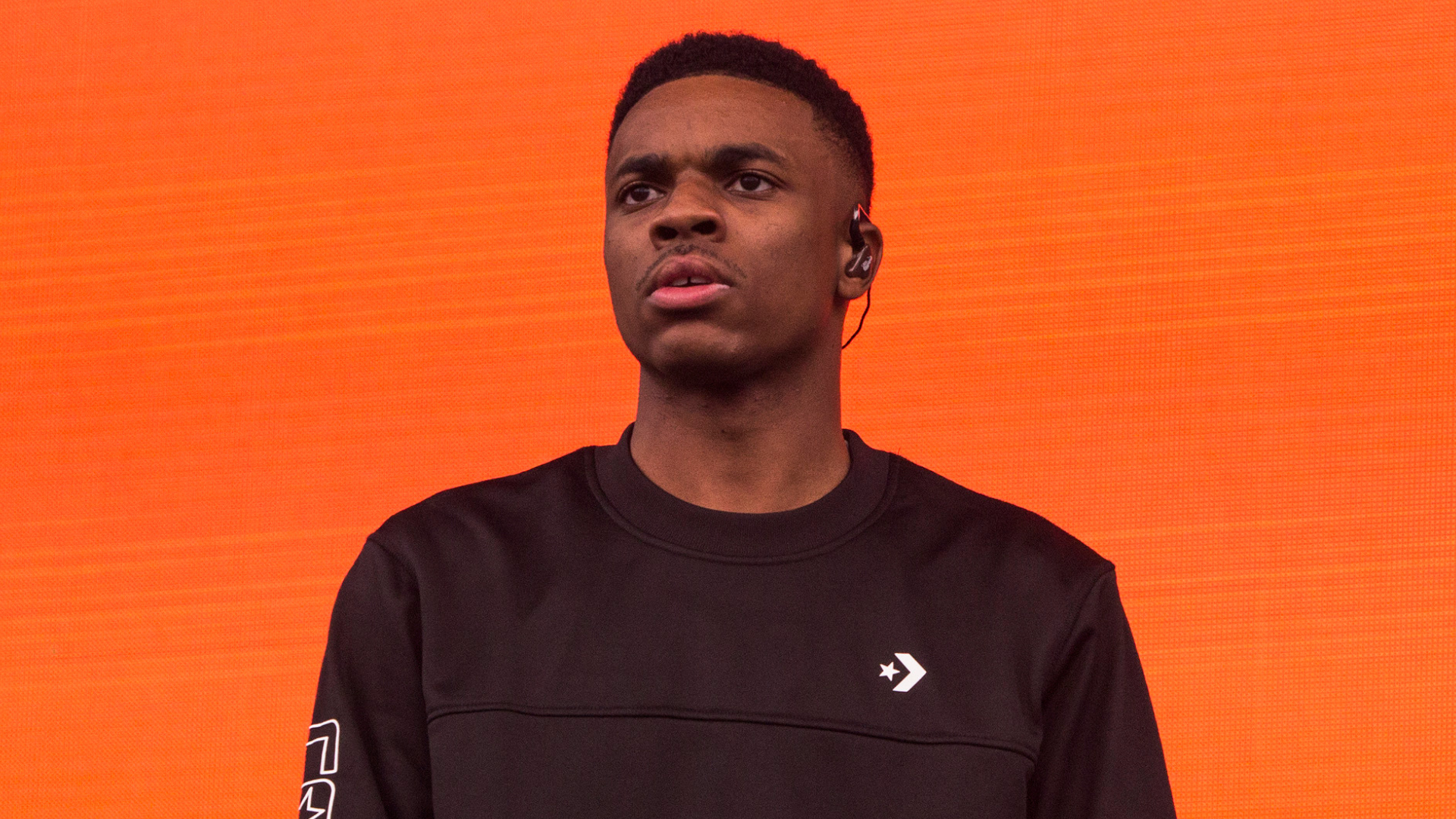 A person wearing a black sweatshirt with an orange background, looking forward with an earpiece.