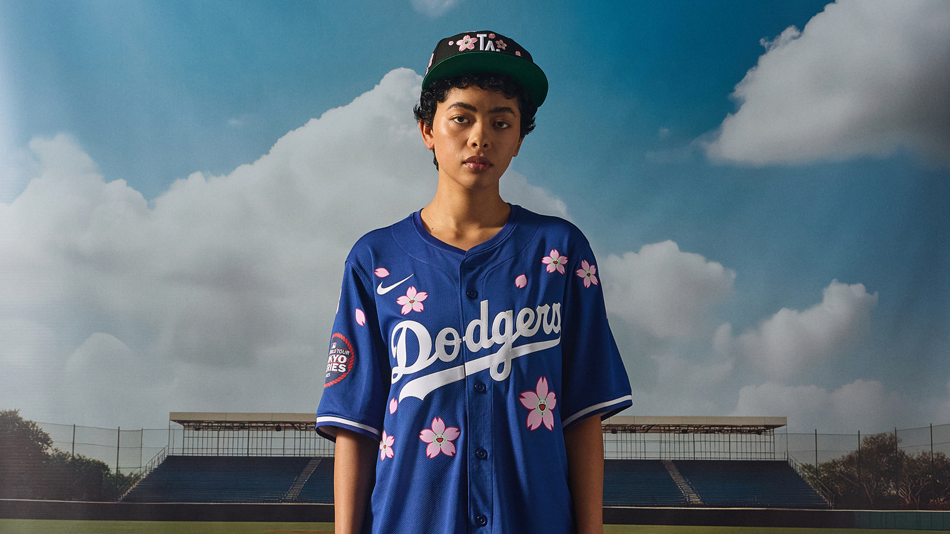 A person wearing a Dodgers jersey with pink flowers and a matching cap stands in front of a baseball field backdrop.