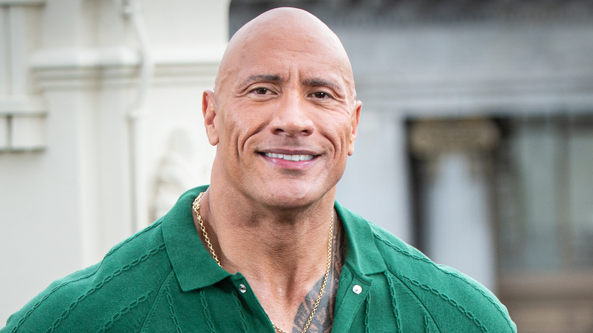 Dwayne "The Rock" Johnson smiling, wearing a green shirt, standing outdoors.