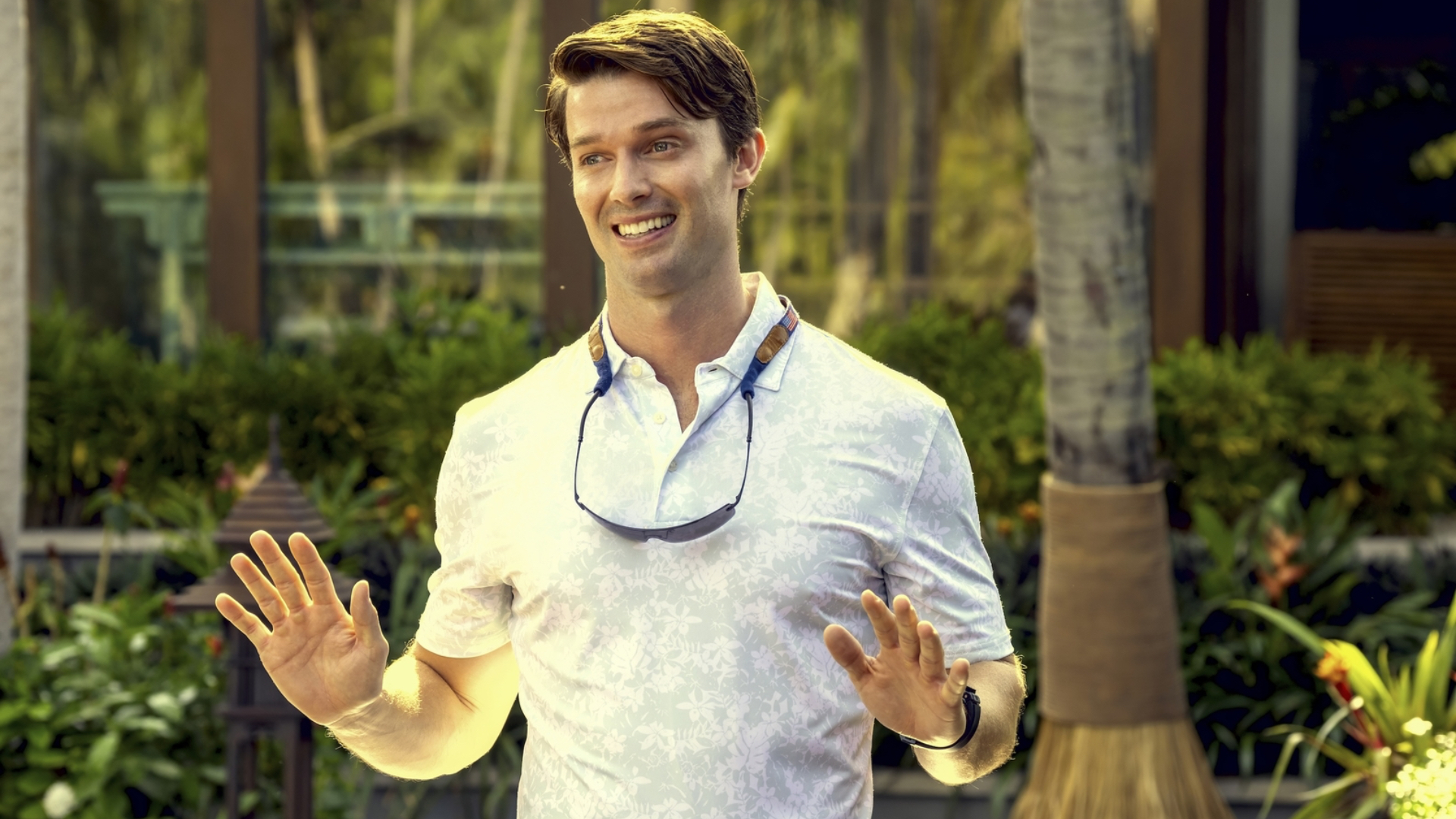 A man in a casual white shirt with sunglasses around his neck, standing outdoors with greenery in the background, smiling and gesturing with his hands.