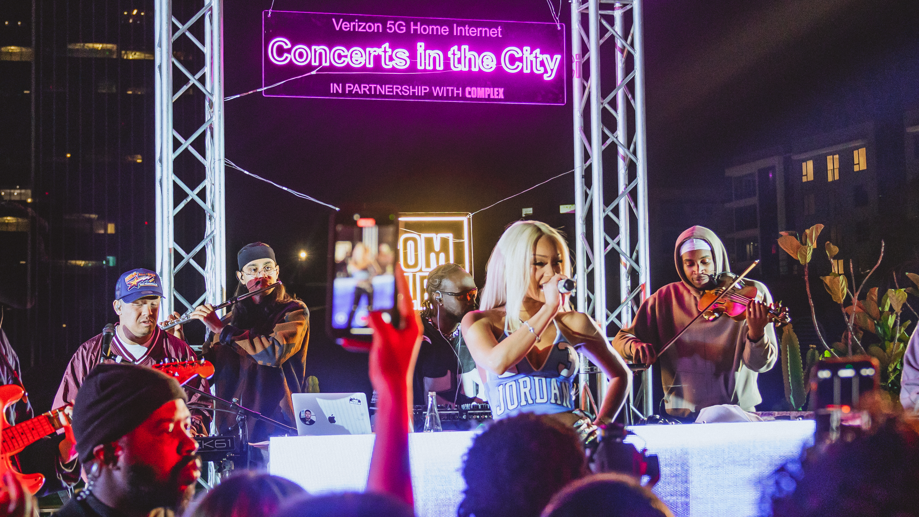 A lively concert scene with a female singer performing on stage, surrounded by musicians. A neon sign reads "Concerts in the City."