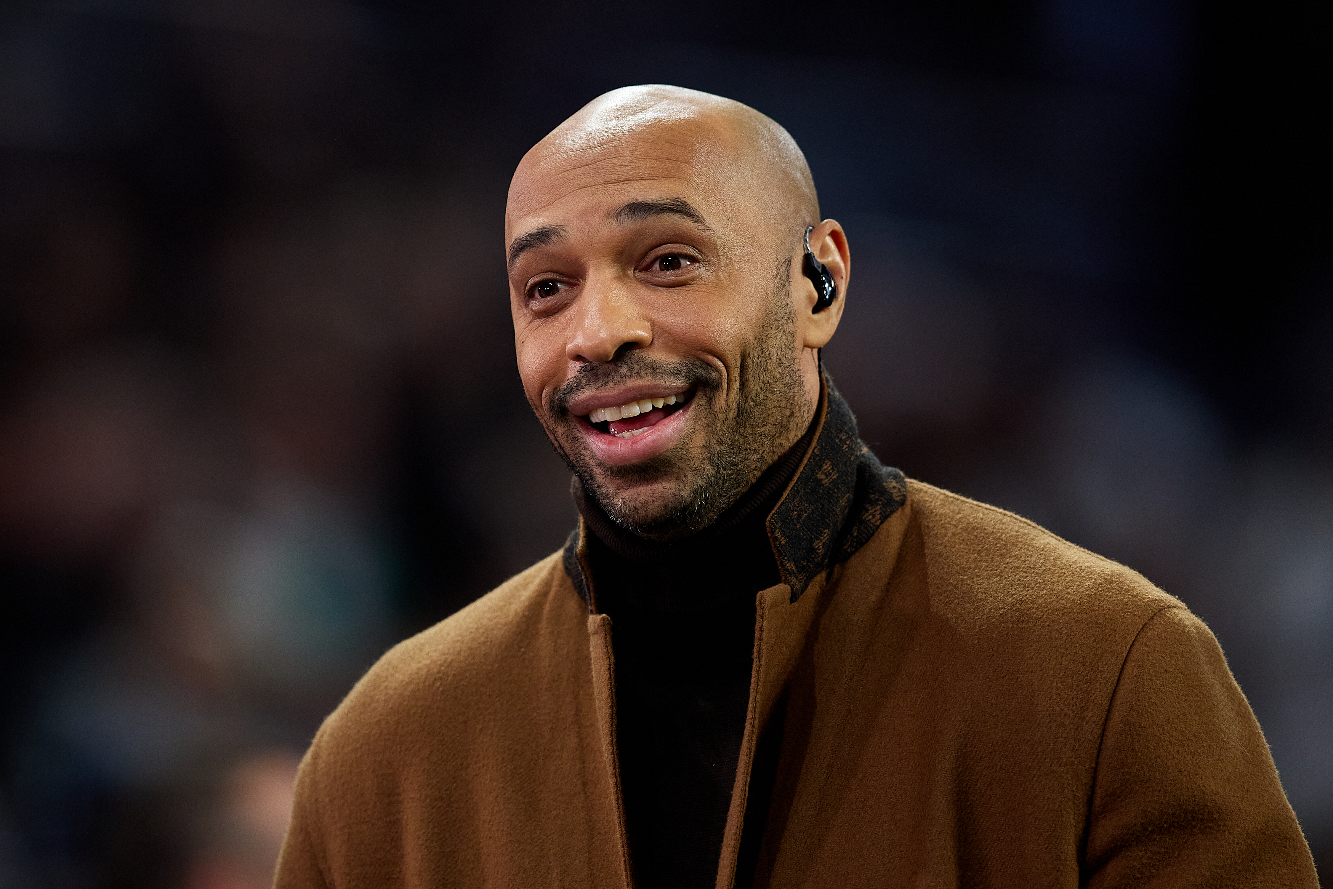 Thierry Henry, wearing a brown coat and earpiece, with a blurred background.