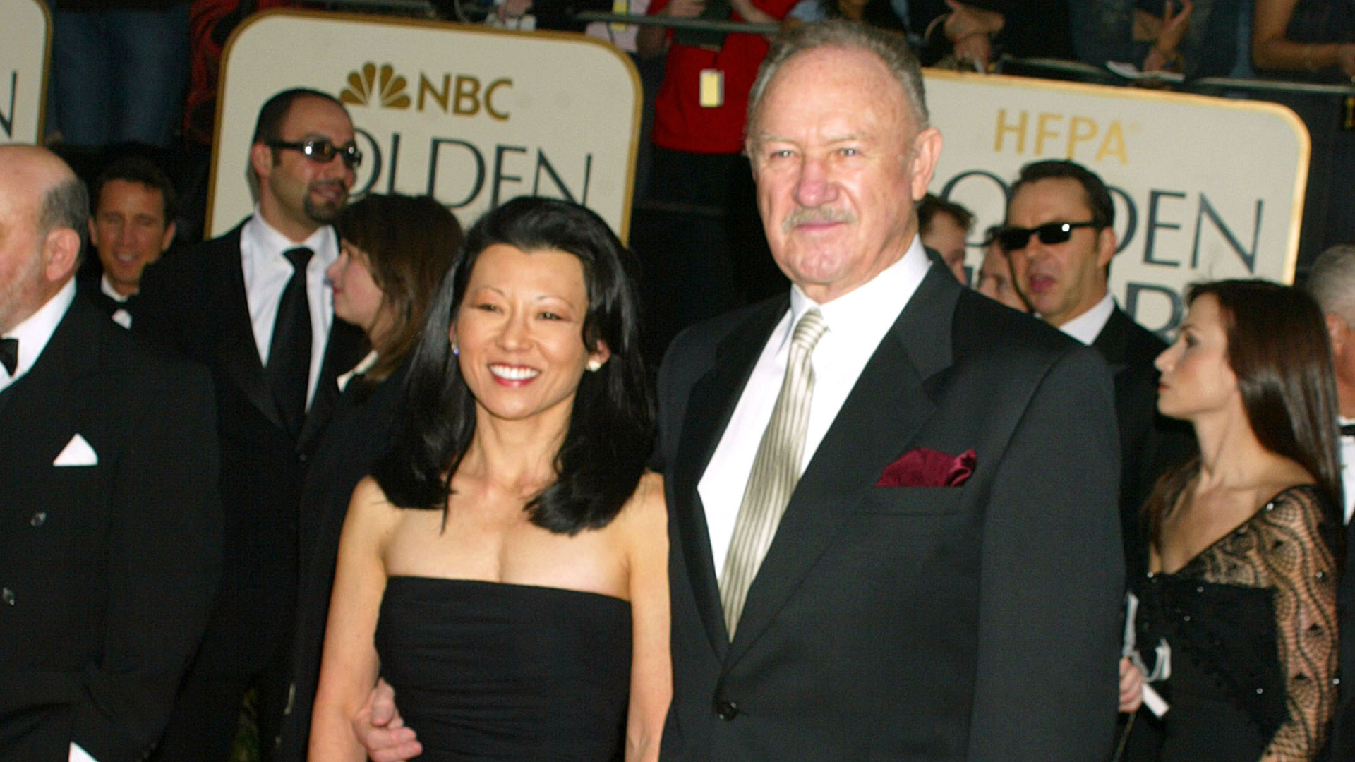 A couple posing at the Golden Globe Awards, with the man in a black suit and the woman in a black dress, surrounded by other attendees.