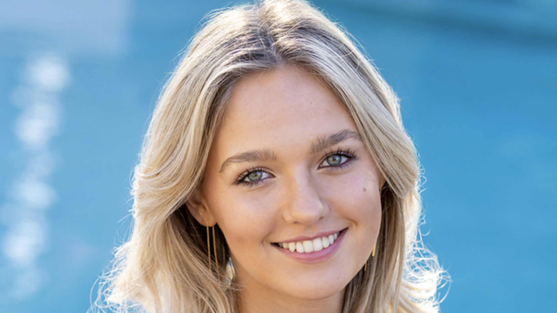 A person with long blonde hair smiles, standing in front of a blurred blue background.