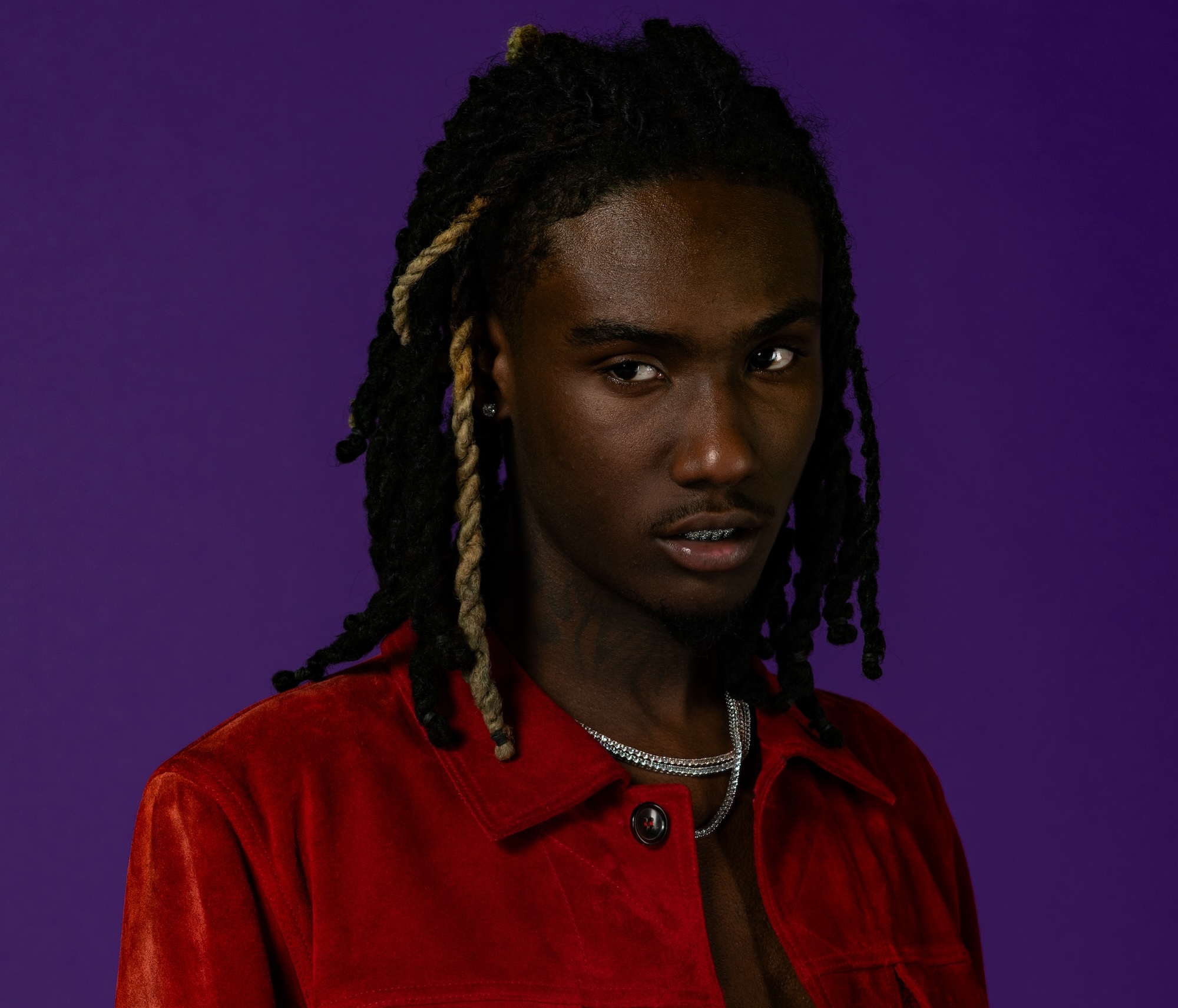 OsamaSon dreadlocks, wearing a red jacket and a chain necklace, poses against a purple background.