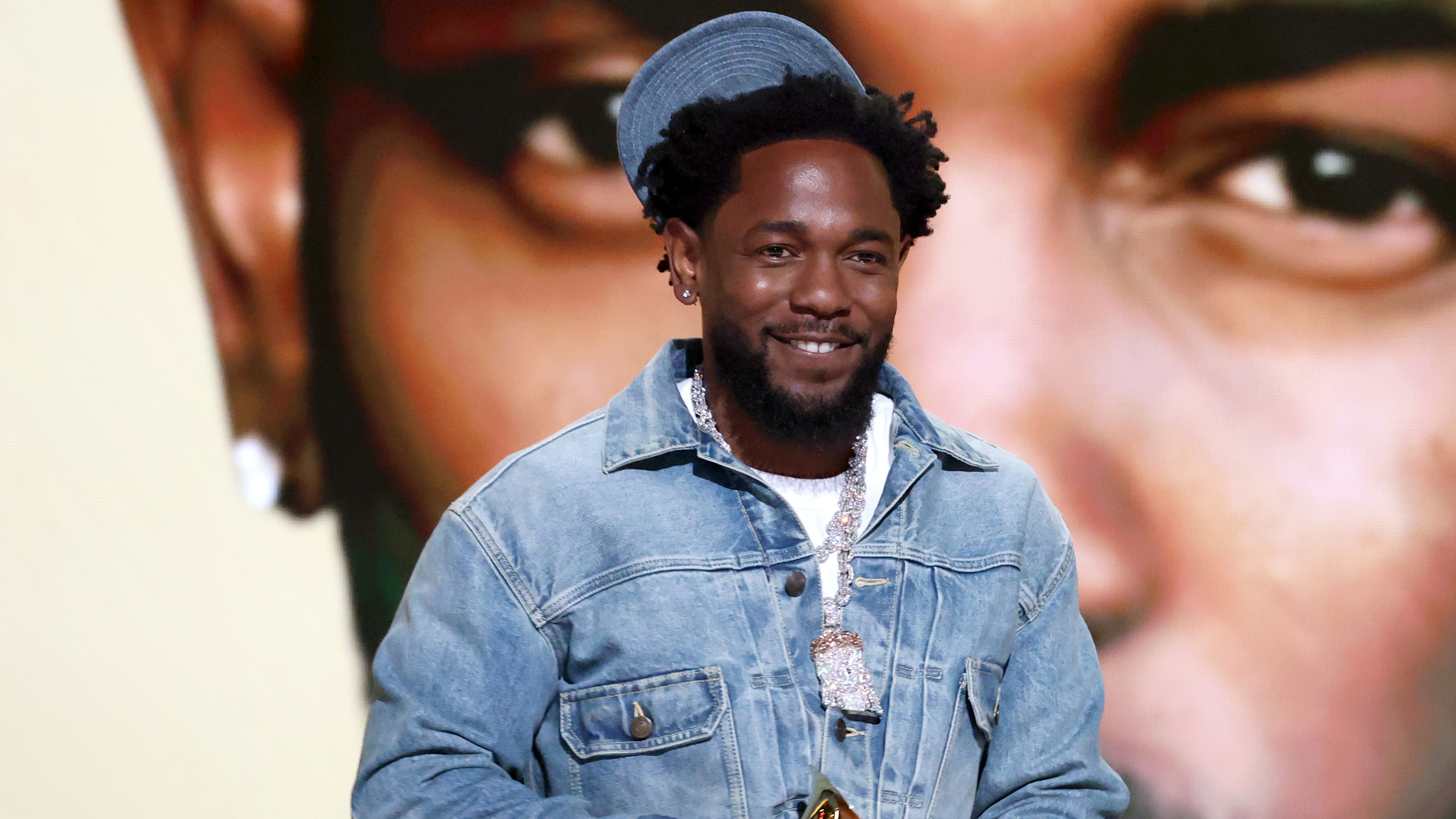 Kendrick Lamar, wearing a denim jacket and hat, smiles while holding a Grammy award. A large image of his face is in the background.