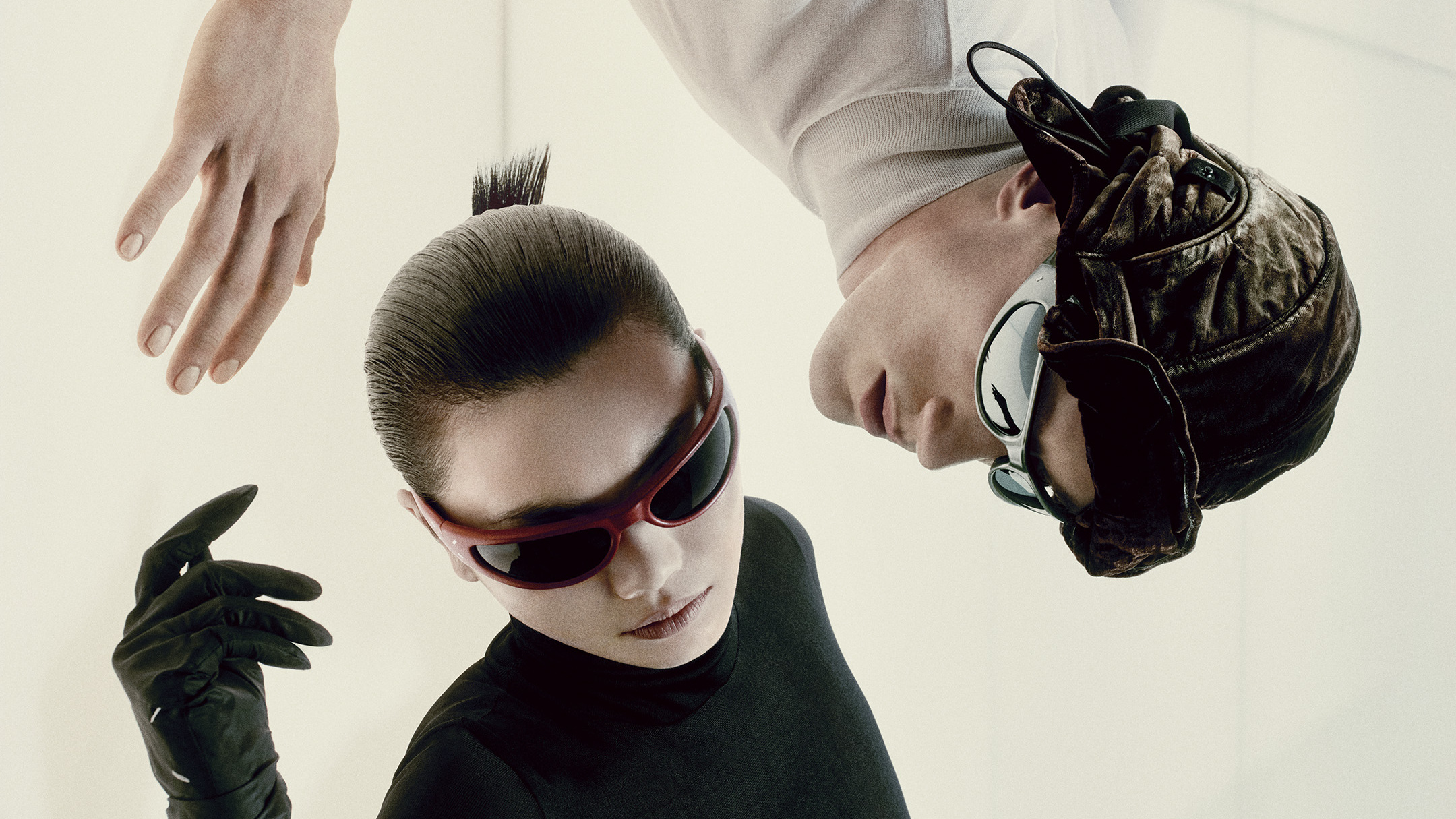 Two individuals wearing futuristic sunglasses and stylish outfits pose artistically, one upside down. The setting is minimalist and modern.
