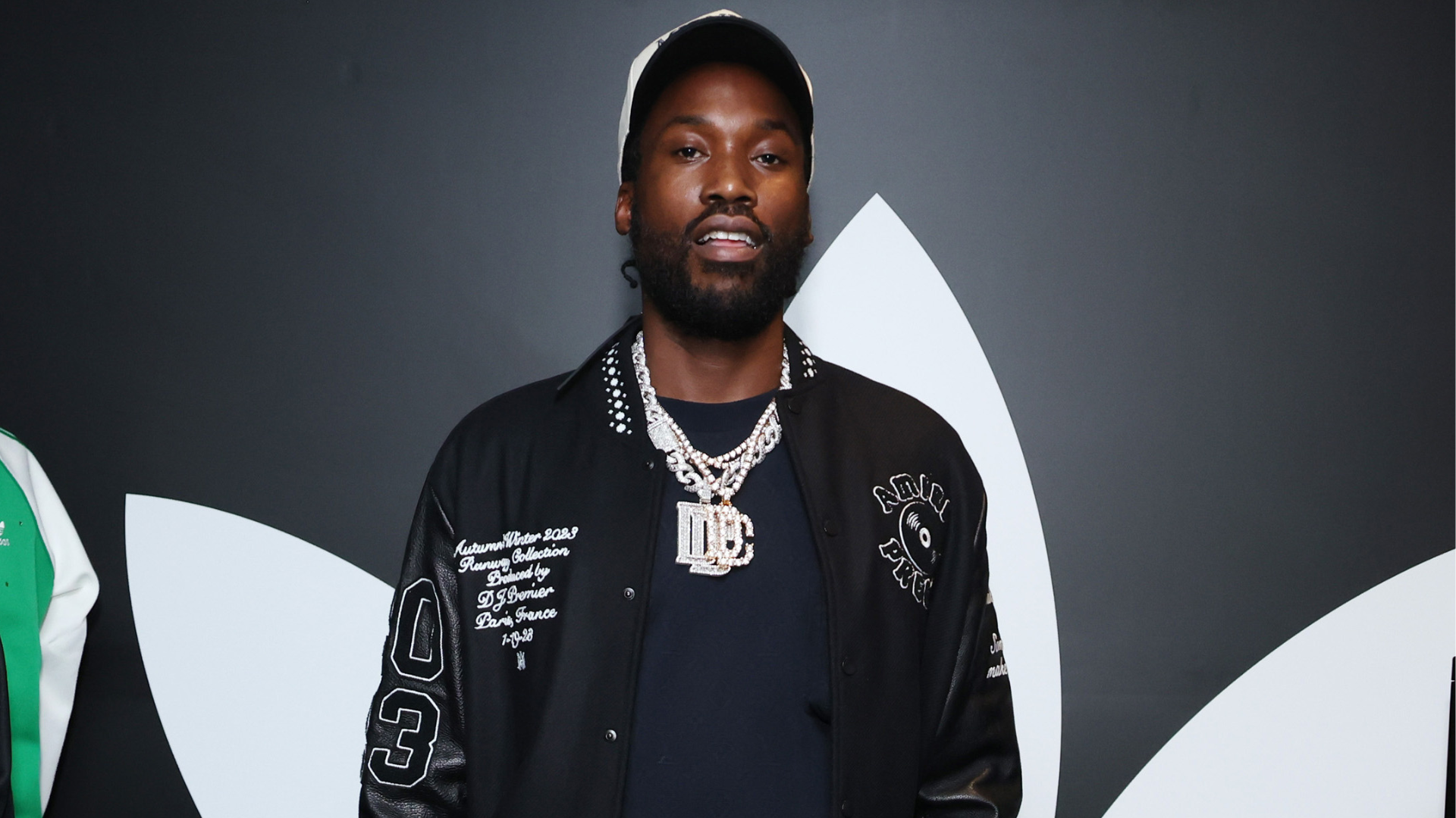 A man wearing a black jacket with a large silver chain necklace, standing in front of a dark background with a white design.