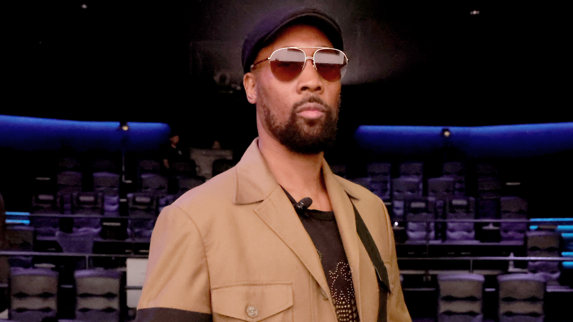 A man wearing sunglasses, a black cap, and a tan jacket stands in a dimly lit theater with empty seats in the background.