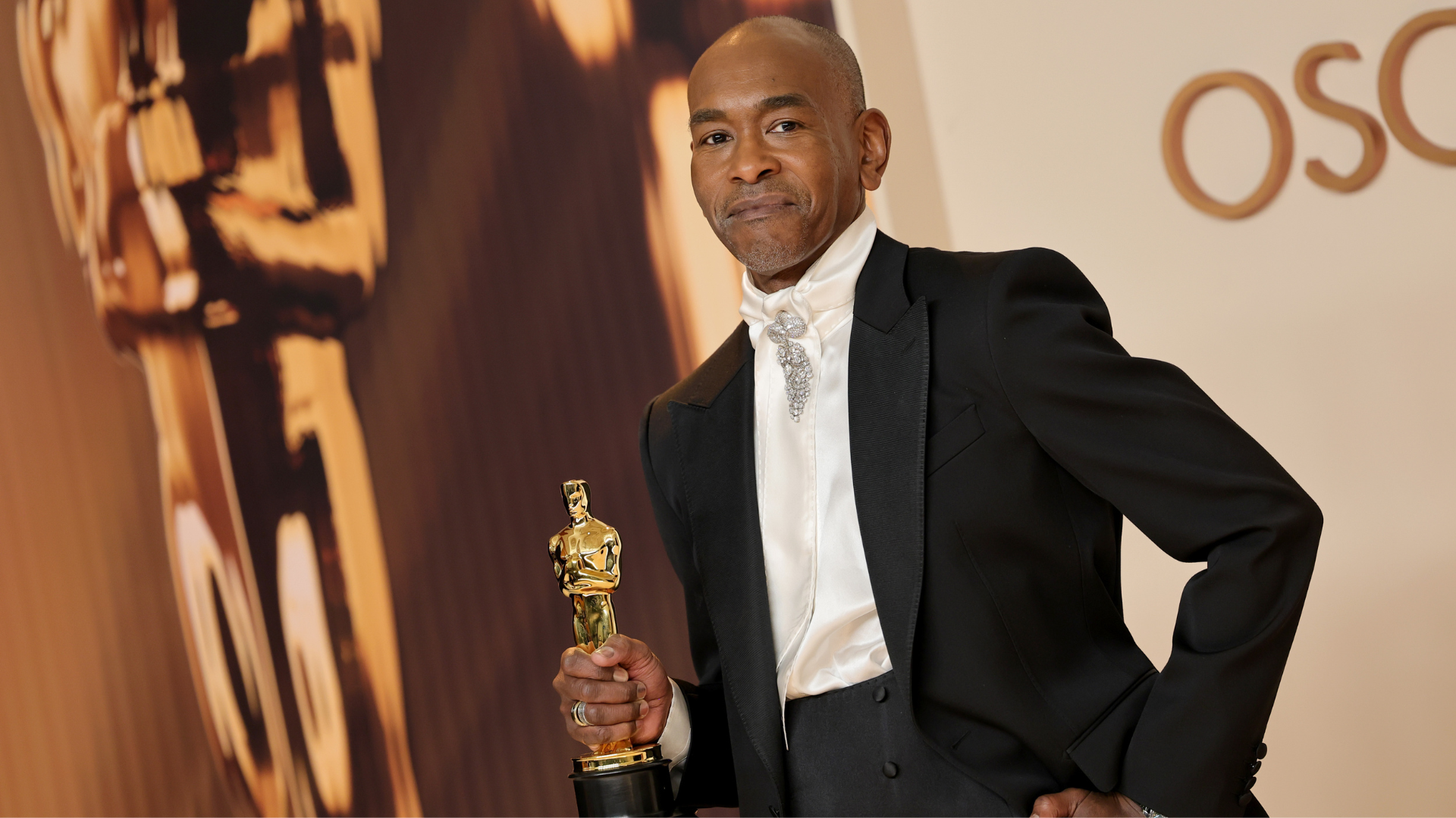 A man in formal attire holding an Oscar statuette, standing in front of a backdrop with the word "Oscar" partially visible.