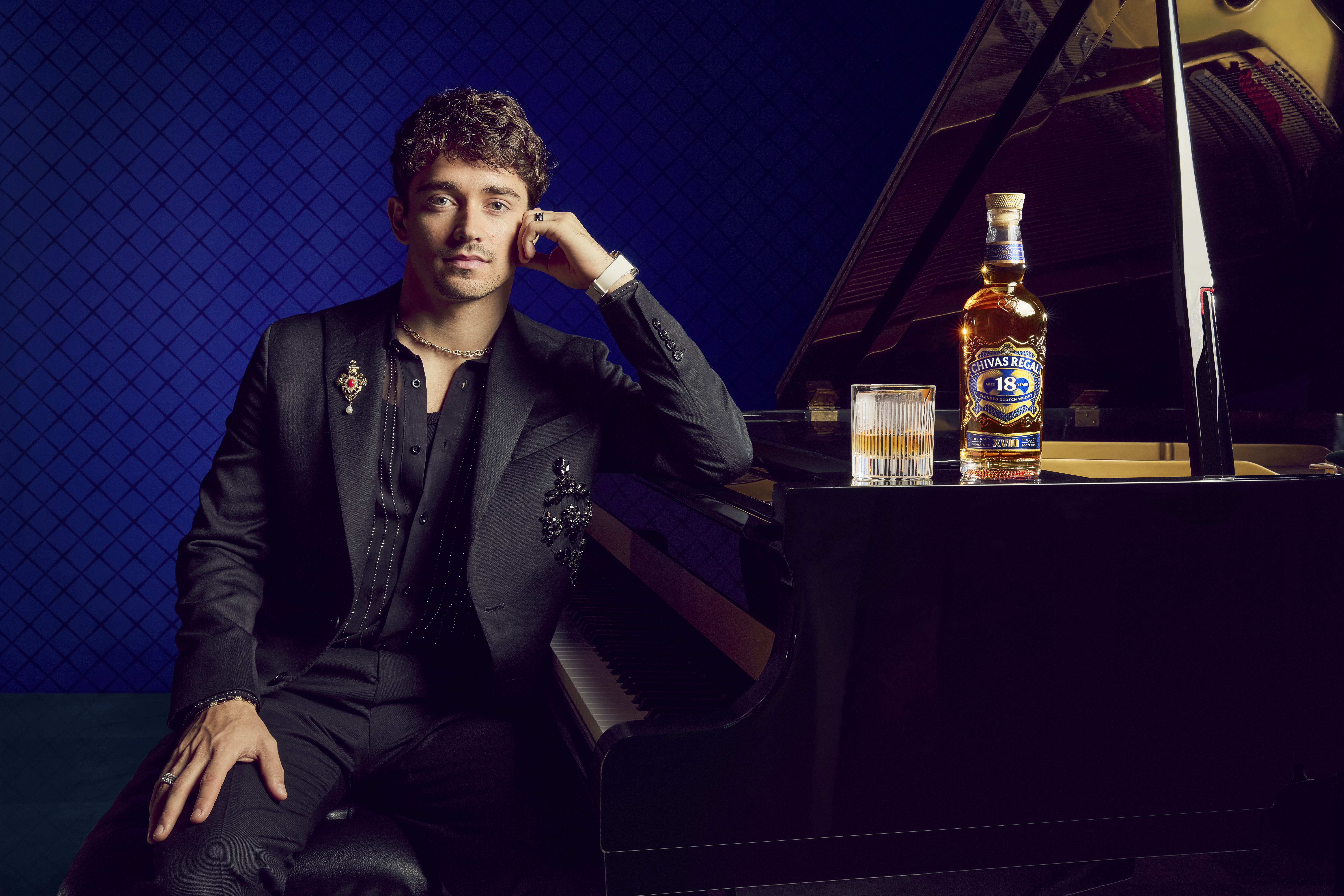 A man in a black suit sits at a grand piano with a bottle of Chivas Regal 18 and a glass. The background is a dark blue pattern.