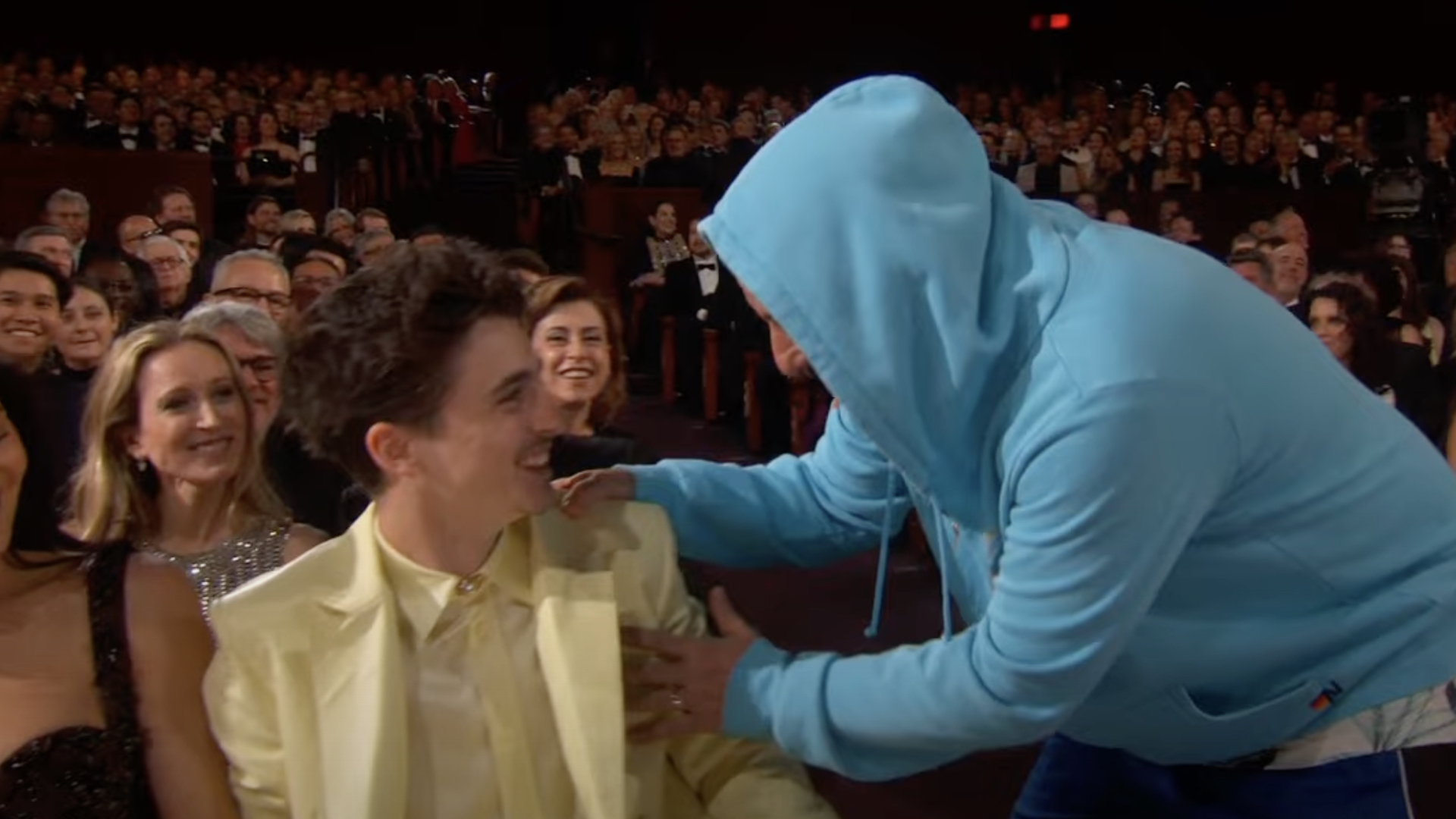A person in a blue hoodie interacts with Timothée Chalamet, who is smiling and wearing a yellow suit, in a crowded auditorium.