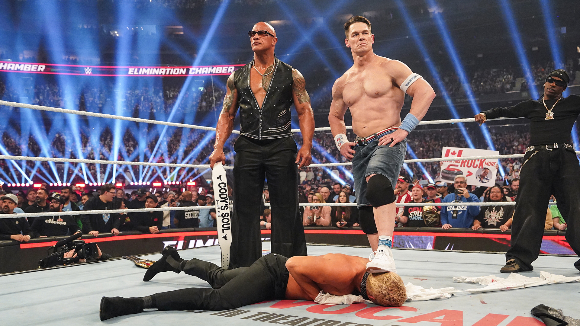 Dwayne "The Rock" Johnson, John Cena, and Travis Scott stand over Cody Rhodes during WWE Elimination Chamber.